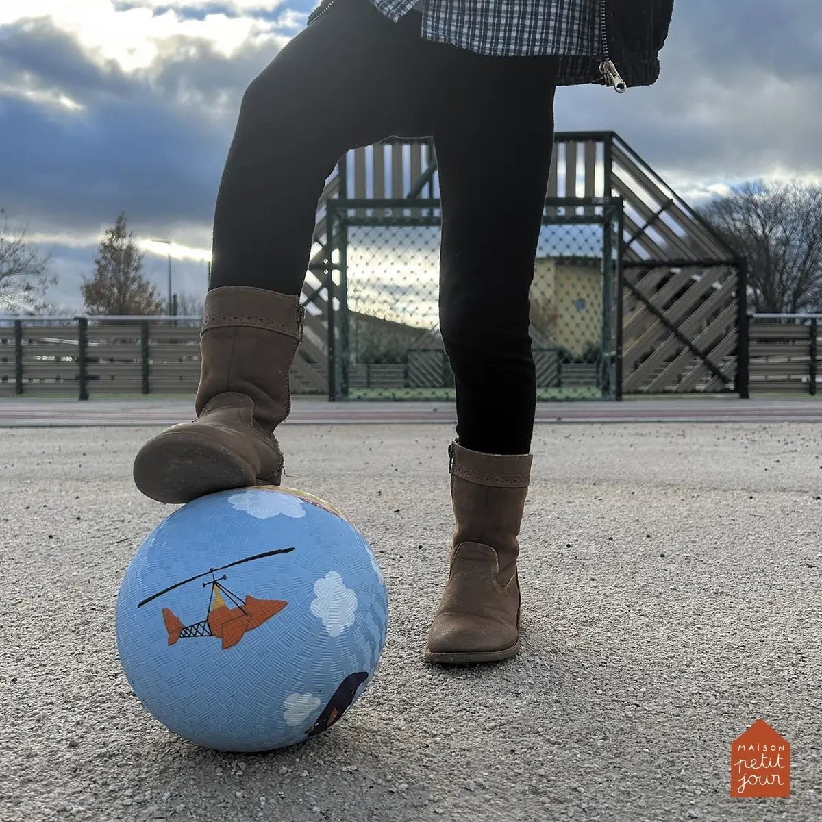 Person wearing brown boots stepping on a blue bouncy  ball with a helicopter design in an outdoor setting.
