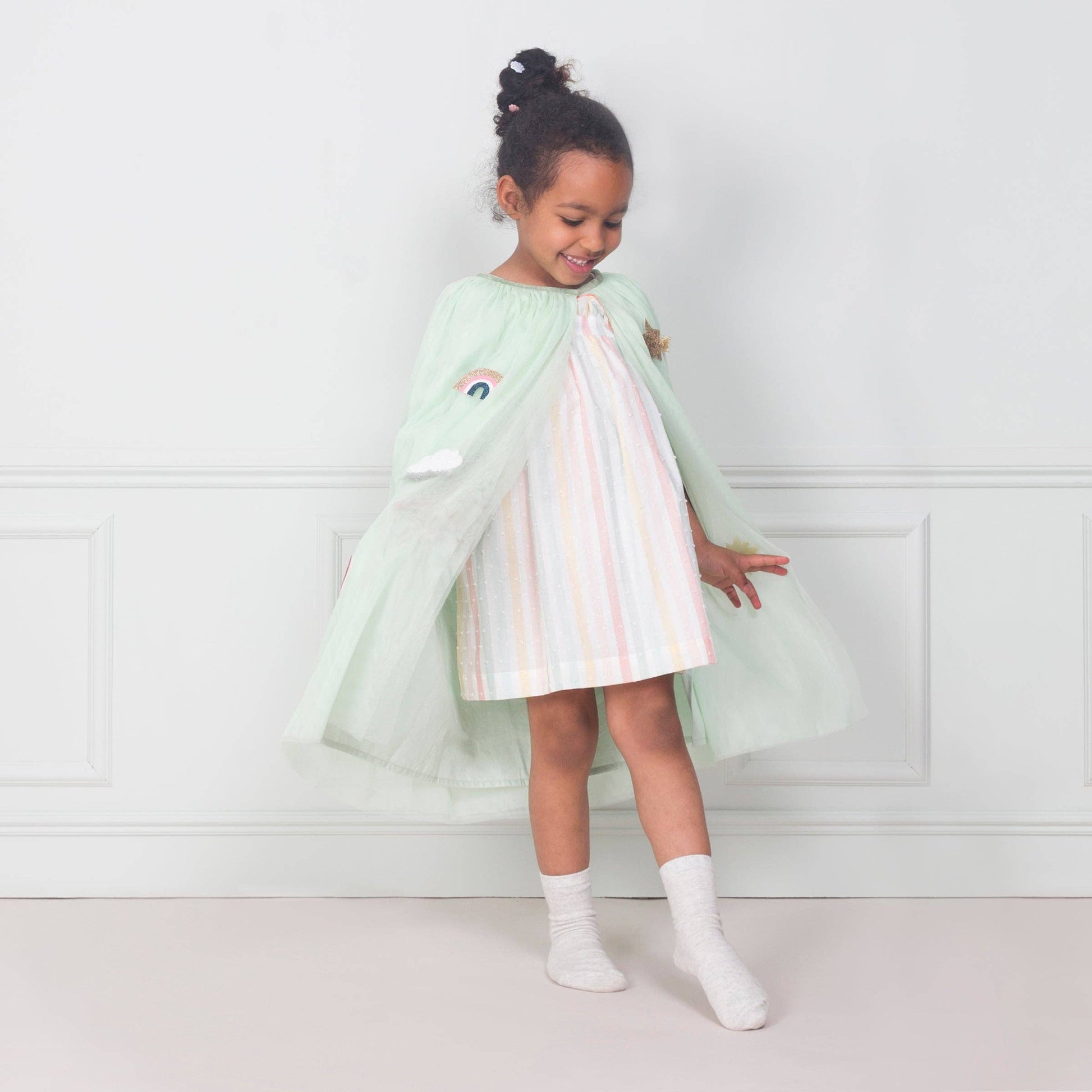Young girl wearing a light green cape with a rainbow dress, standing against a white wall.