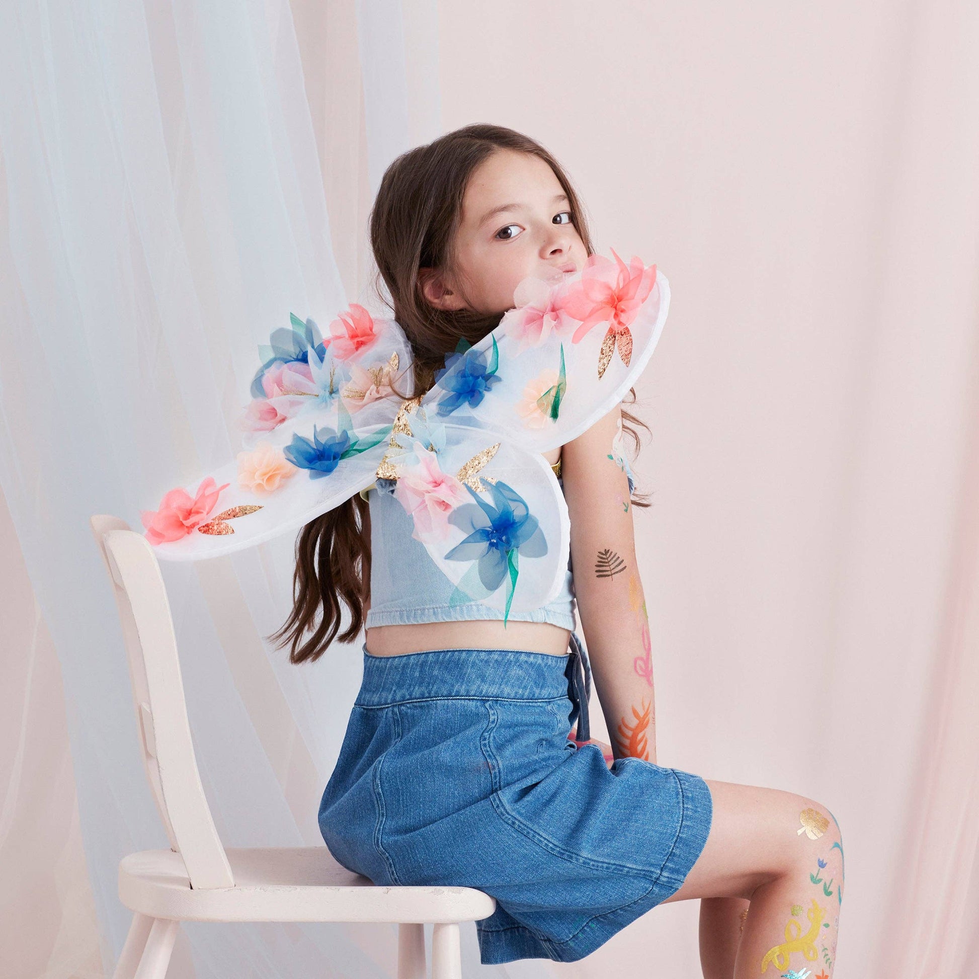 Young girl wearing floral wings sitting on a chair against a light background