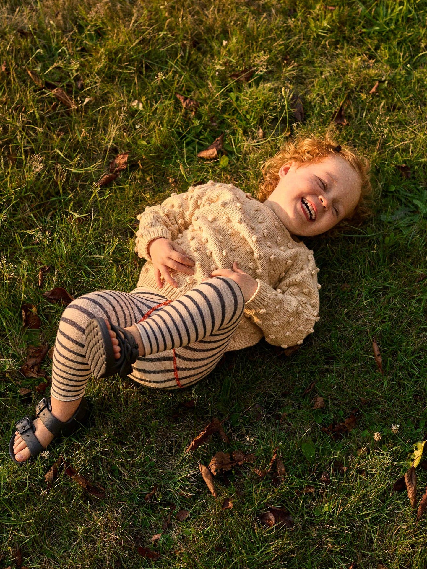 Child lying on grass wearing a beige sweater and striped leggings.