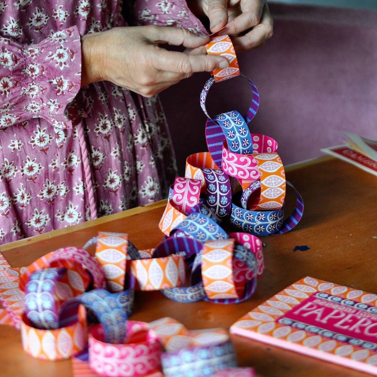 Person assembling colorful Paperchain 4 links on a wooden table beside the patterned kit wallet.