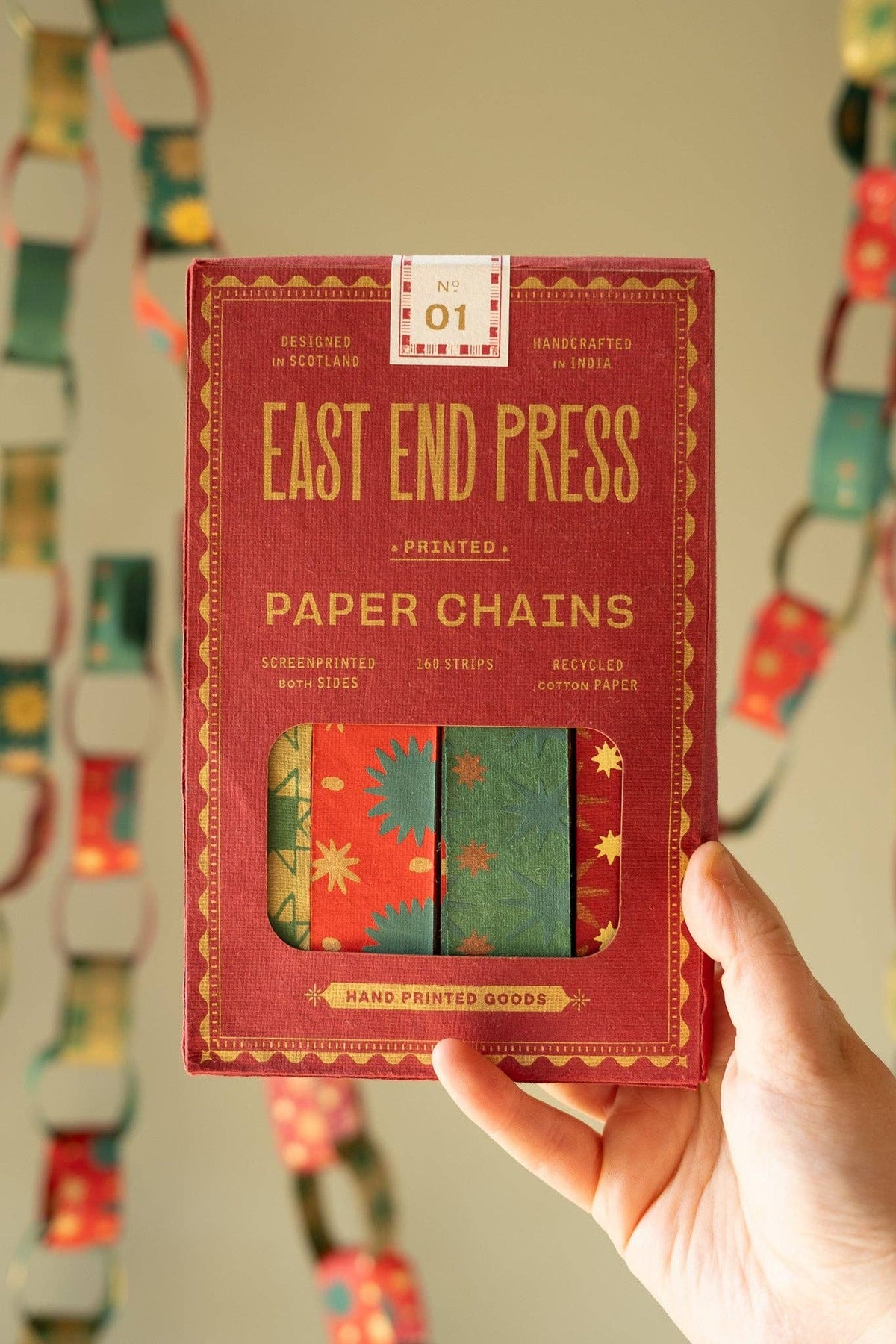 Hand holding the East End Press Festive Paper Chain Kit box in front of hanging paper chains.