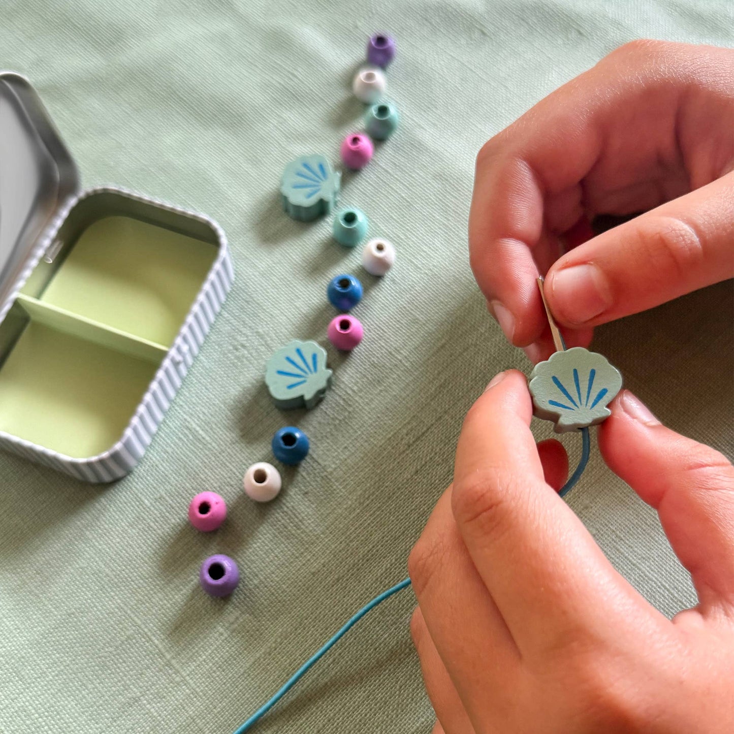 Person stringing colorful beads on a thread with a small container of beads on a light green fabric background.