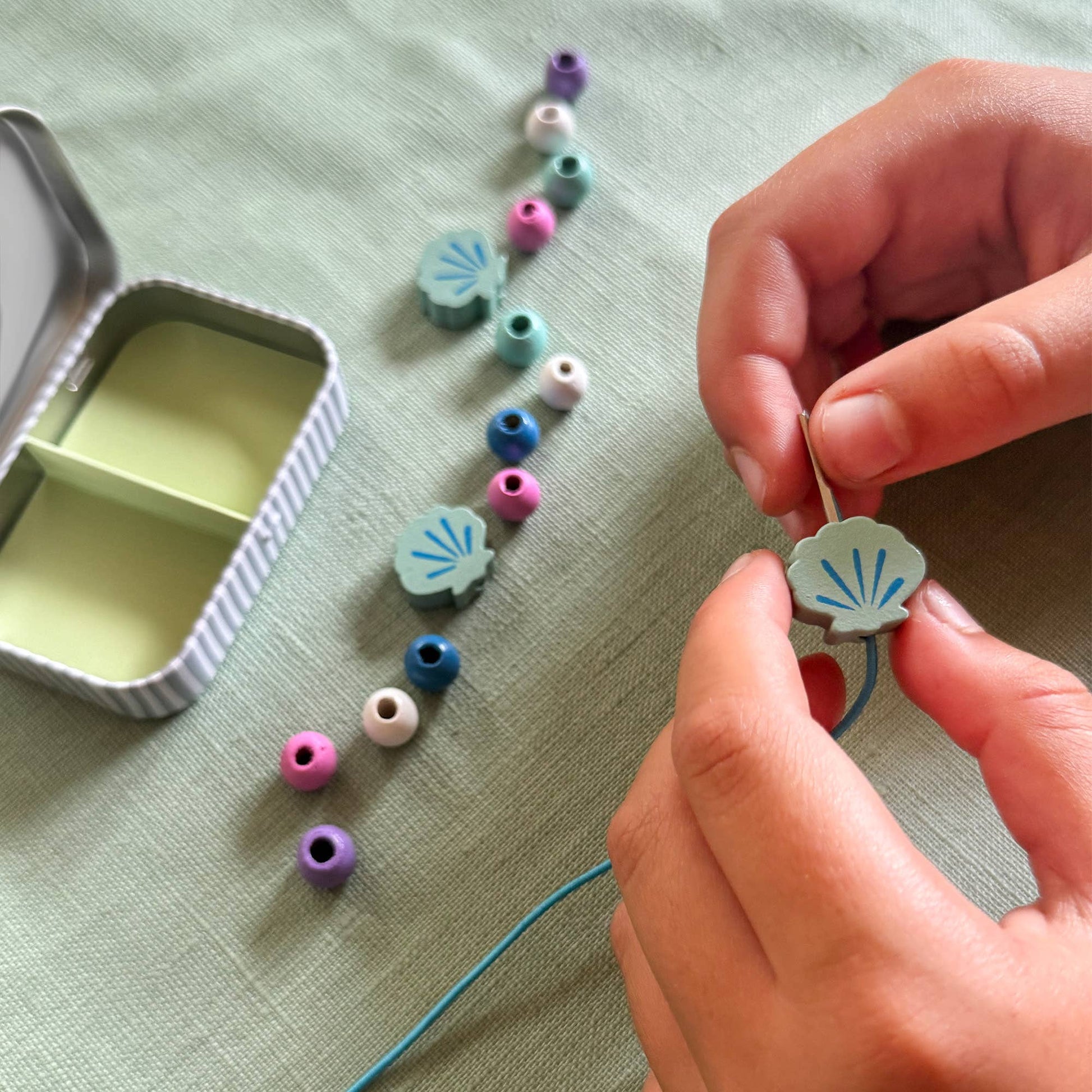 Person stringing colorful beads on a thread with a small container of beads on a light green fabric background.