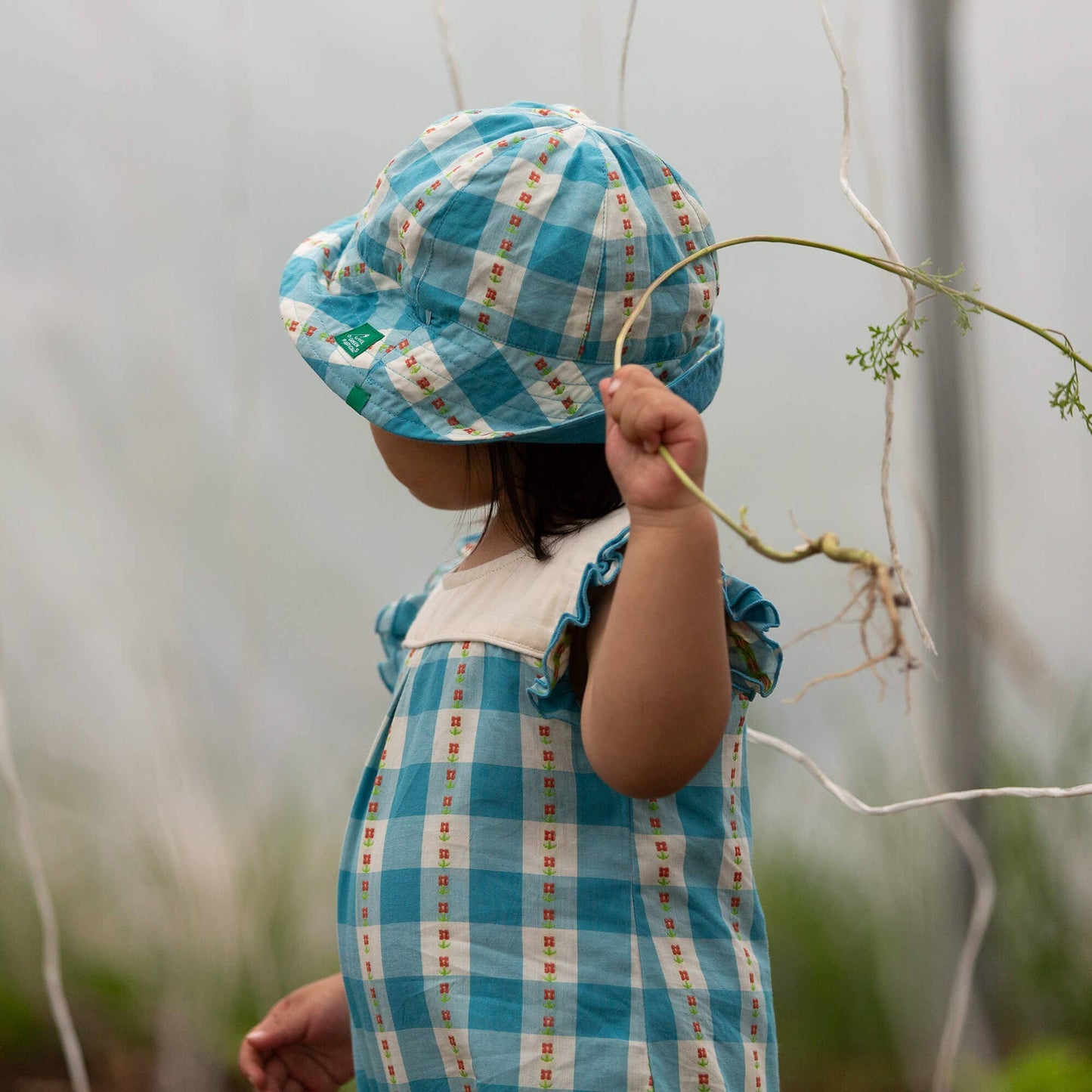 Child wearing a blue checkered hat and dress holding a plant in a garden.