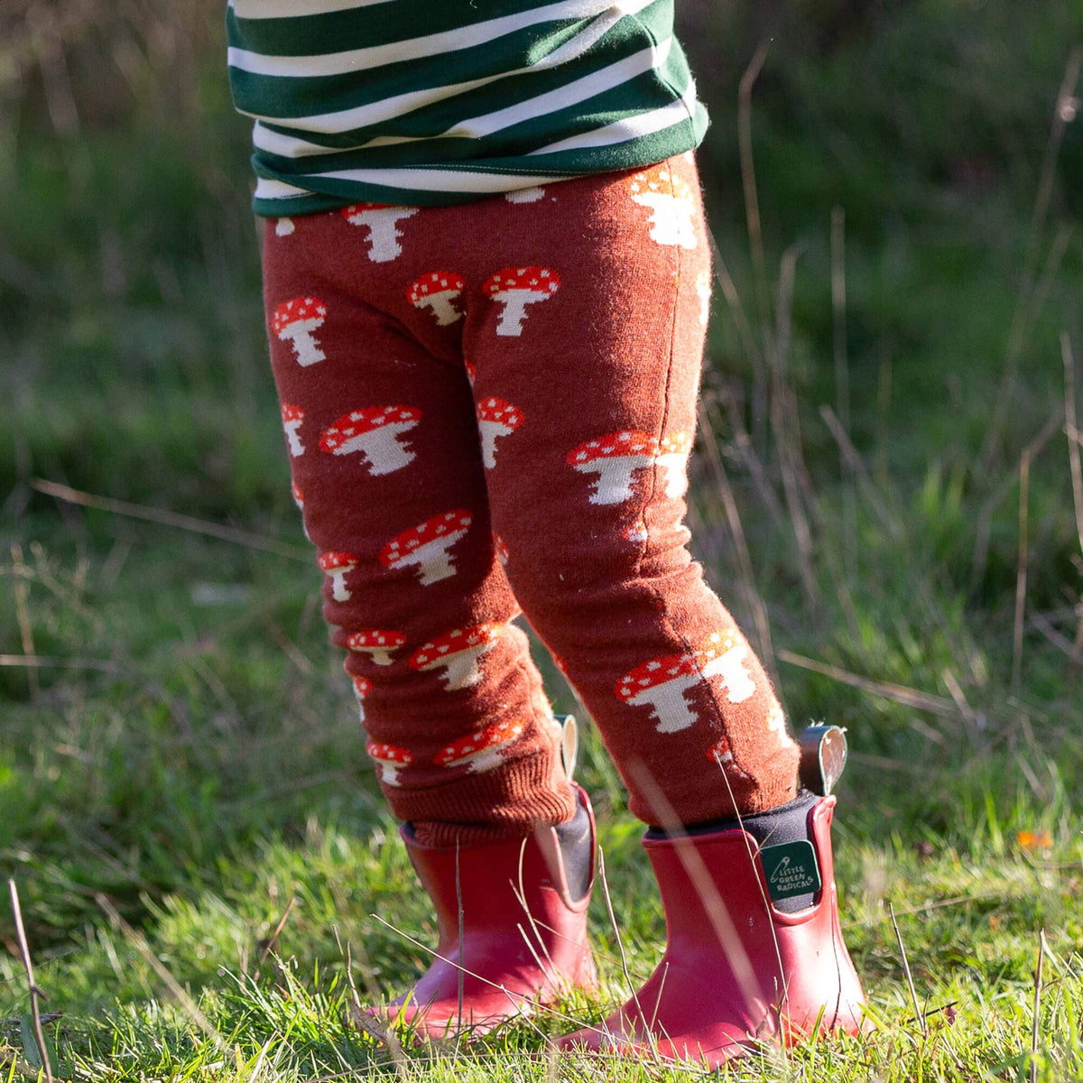 Child wearing rust mushroom-patterned knitted joggers with red rain boots outdoors.
