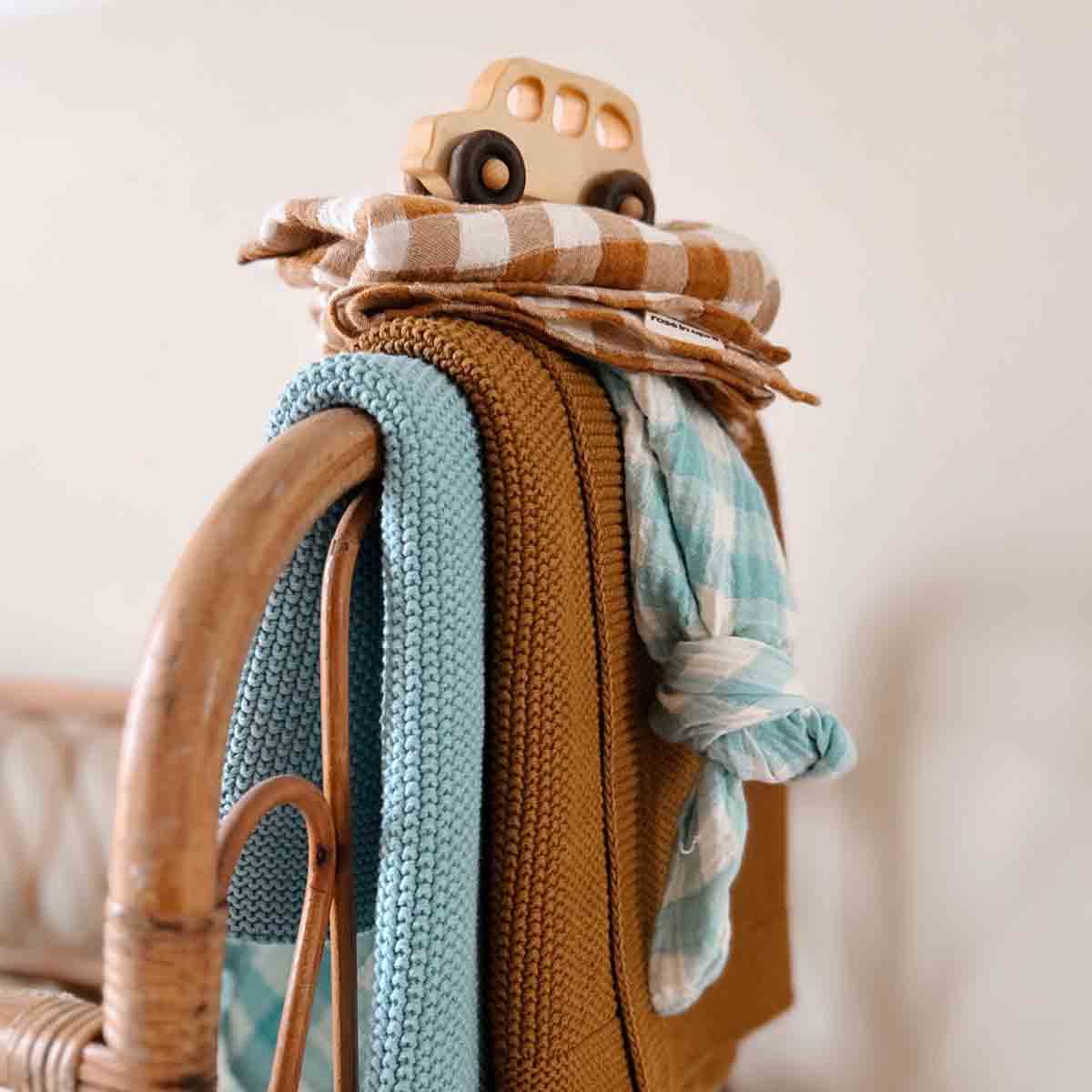 Stack of blankets with a wooden toy car on top, placed on a wicker chair against a neutral background.