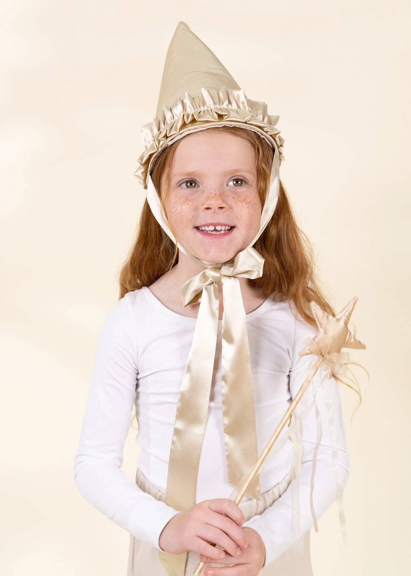 A young girl with freckles wearing a white long-sleeve shirt and a beige pointed wizard hat with a large satin bow, holding a star wand against a solid beige background.
