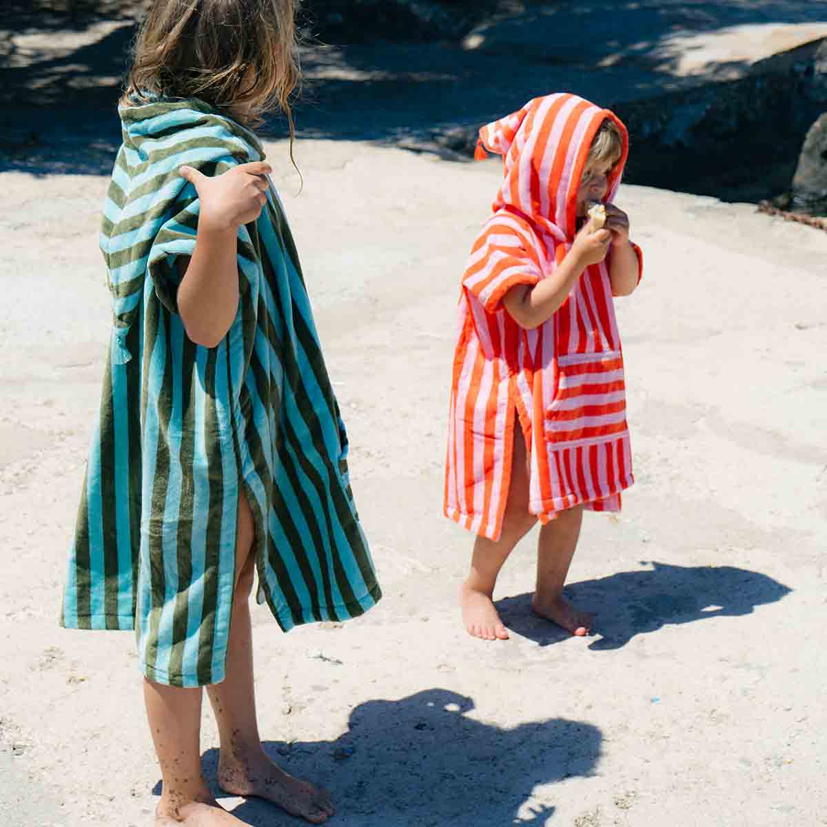 Two children wrapped in striped towels on a sandy beach.