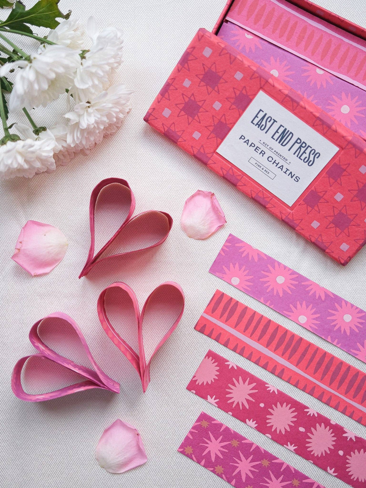 Pink & Red Paper Chain Kit with patterned strips, paper hearts and scattered rose petals on white fabric.