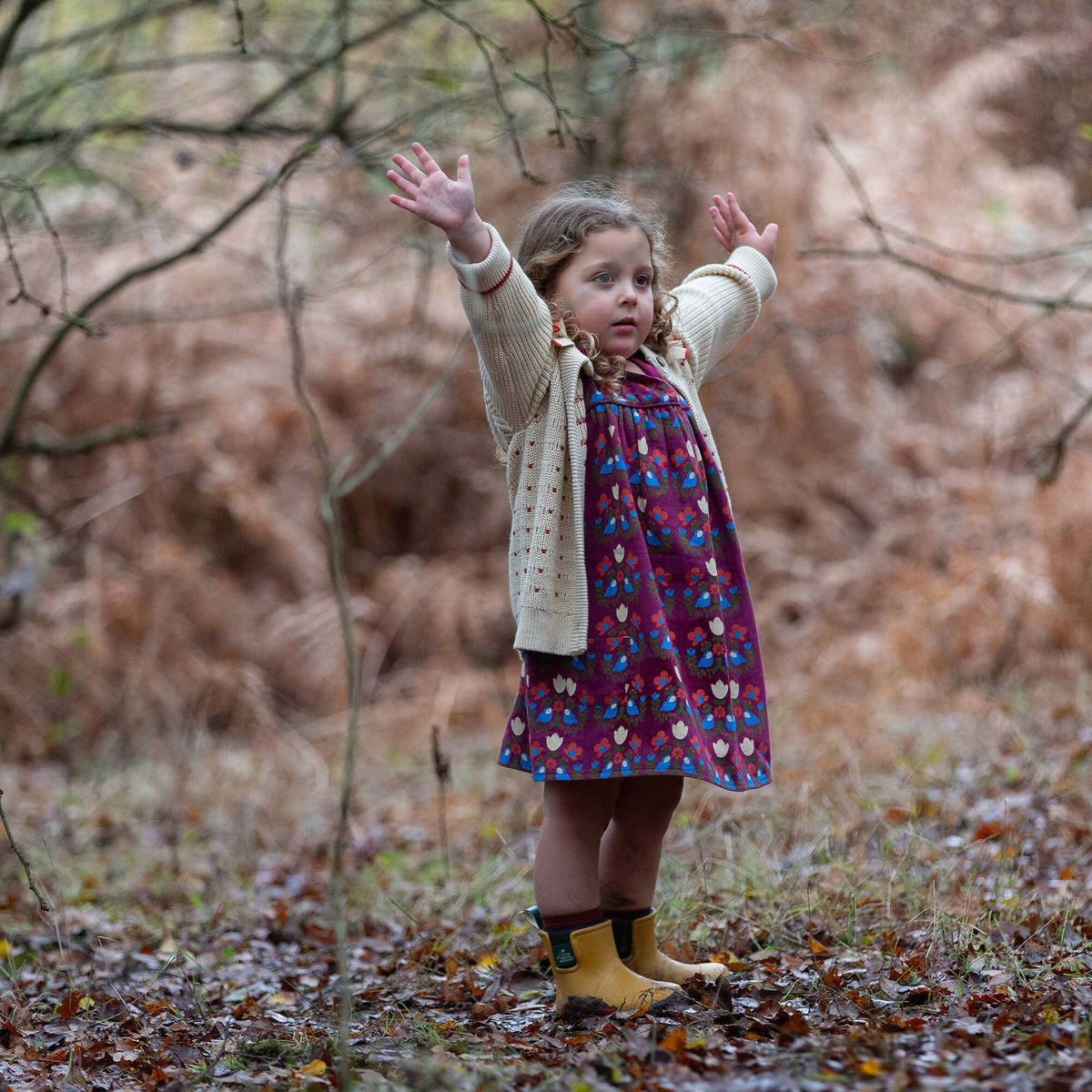 Child wearing a burgundy bird-and-flower swing dress with a cream cardigan in woodland.