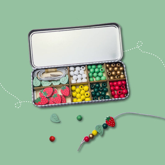 Open tin with compartments of beads and a necklace with leaf and strawberry-shaped beads on a green background