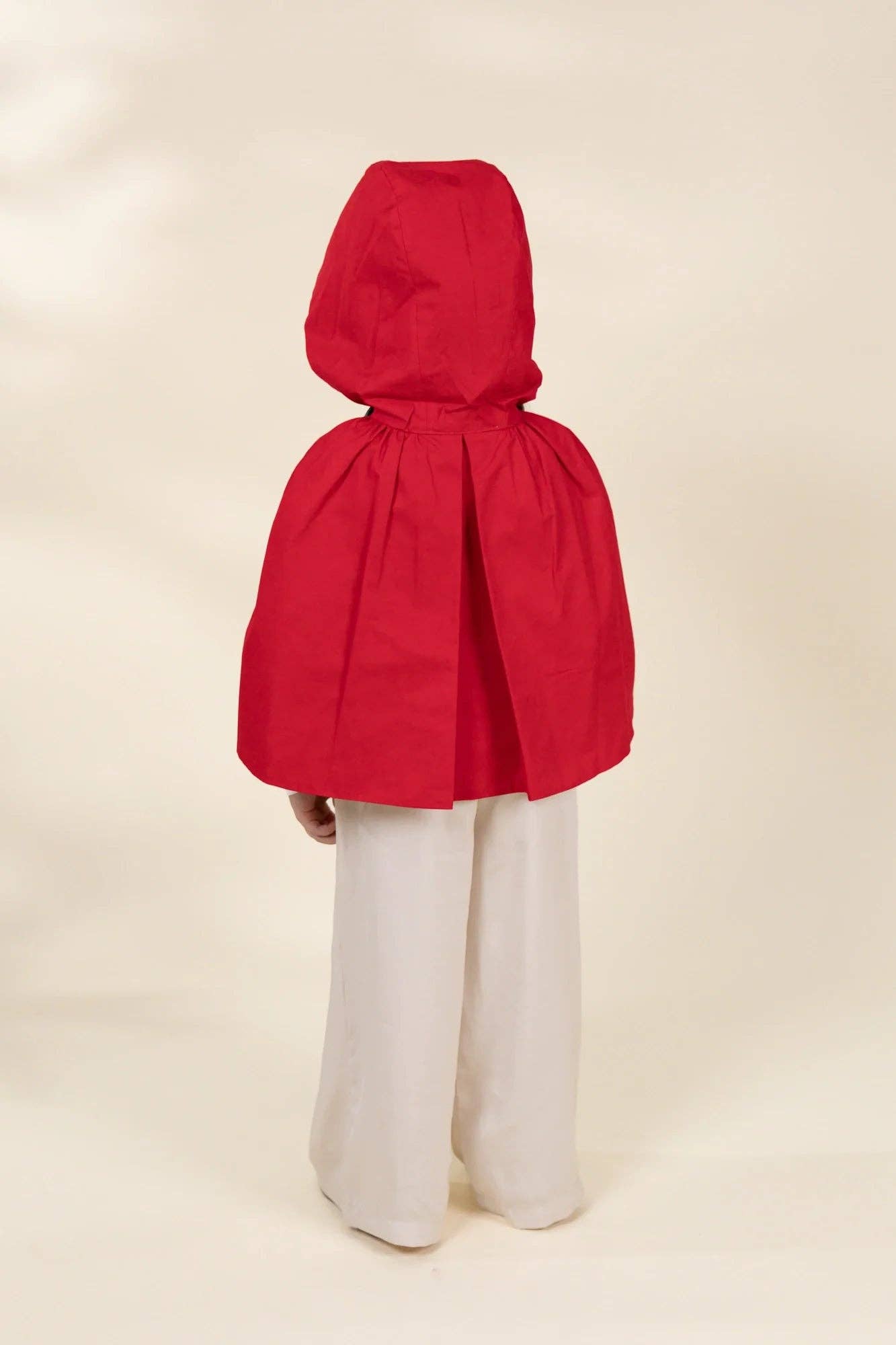 A child wearing a bright red hooded cape with wide-leg light pants, photographed from the back against a plain warm beige studio background.