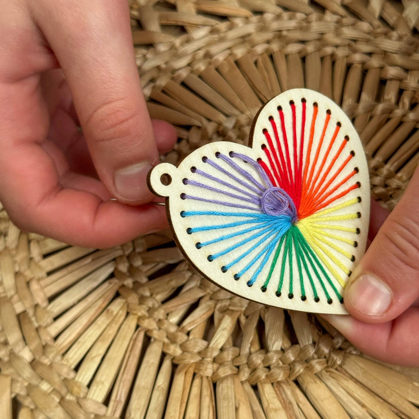 Heart-shaped wooden ornament with colorful string design held by hands on a woven background