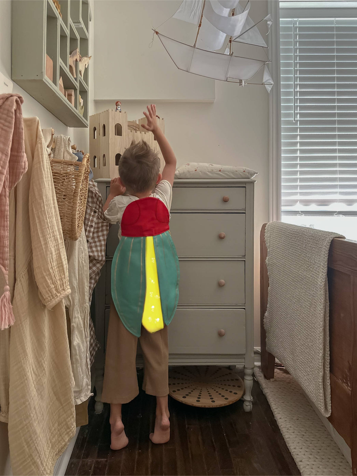Child in a colorful costume standing in a nursery room with a dresser and crib.