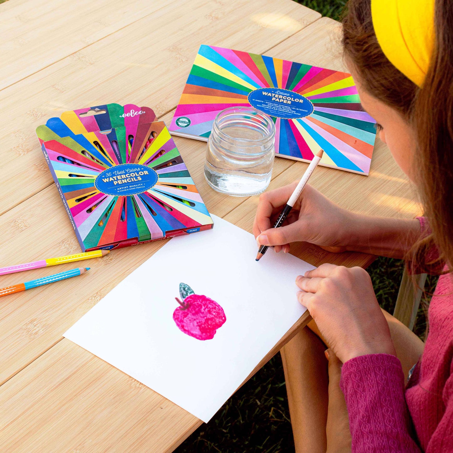 Person drawing with watercolor pencils on a wooden table with a colorful pencil set and water glass.