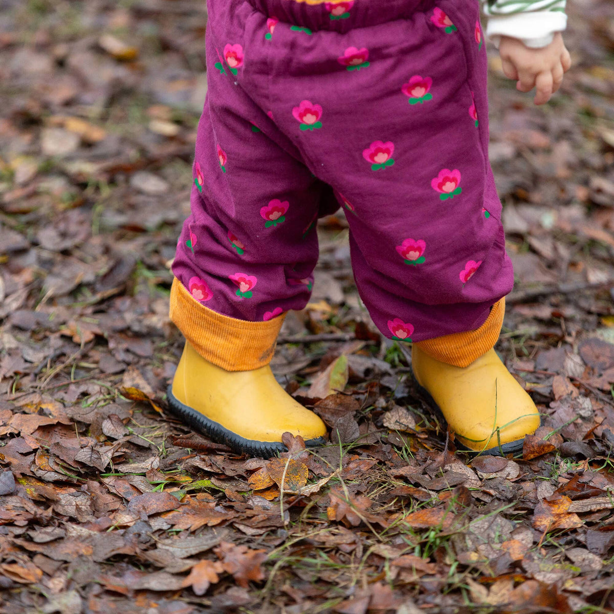 Child wearing purple trousers with pink flowers, mustard turn-ups and yellow rain boots outdoors.
