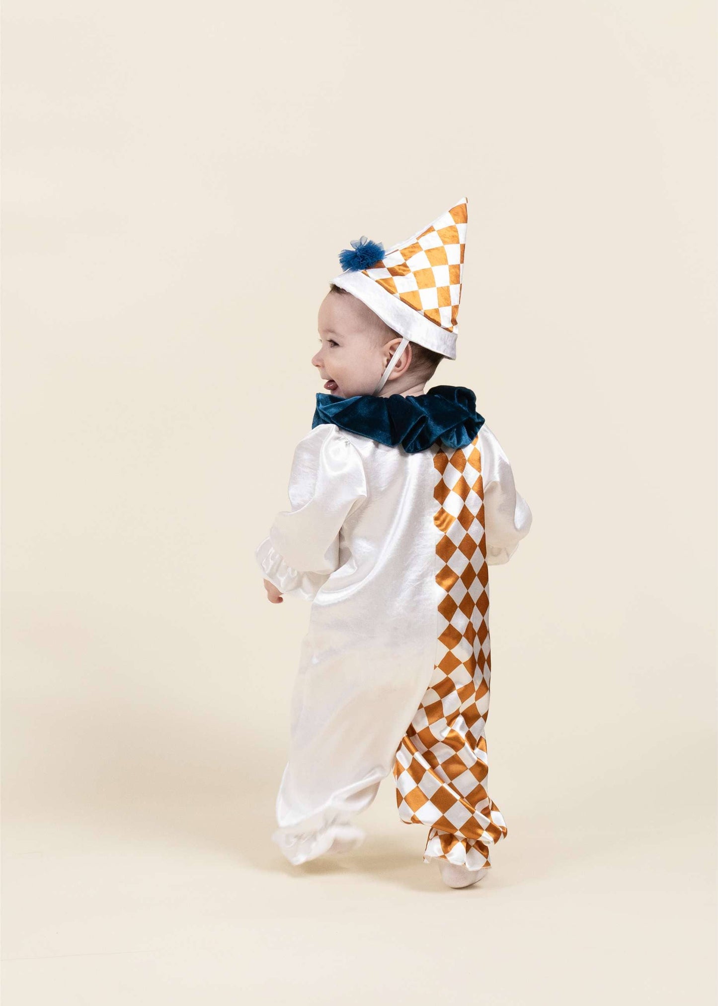A baby wearing a white clown costume with a gold and white checkered pattern on the back and hat, and a dark blue ruffled collar, standing against a solid beige background.
