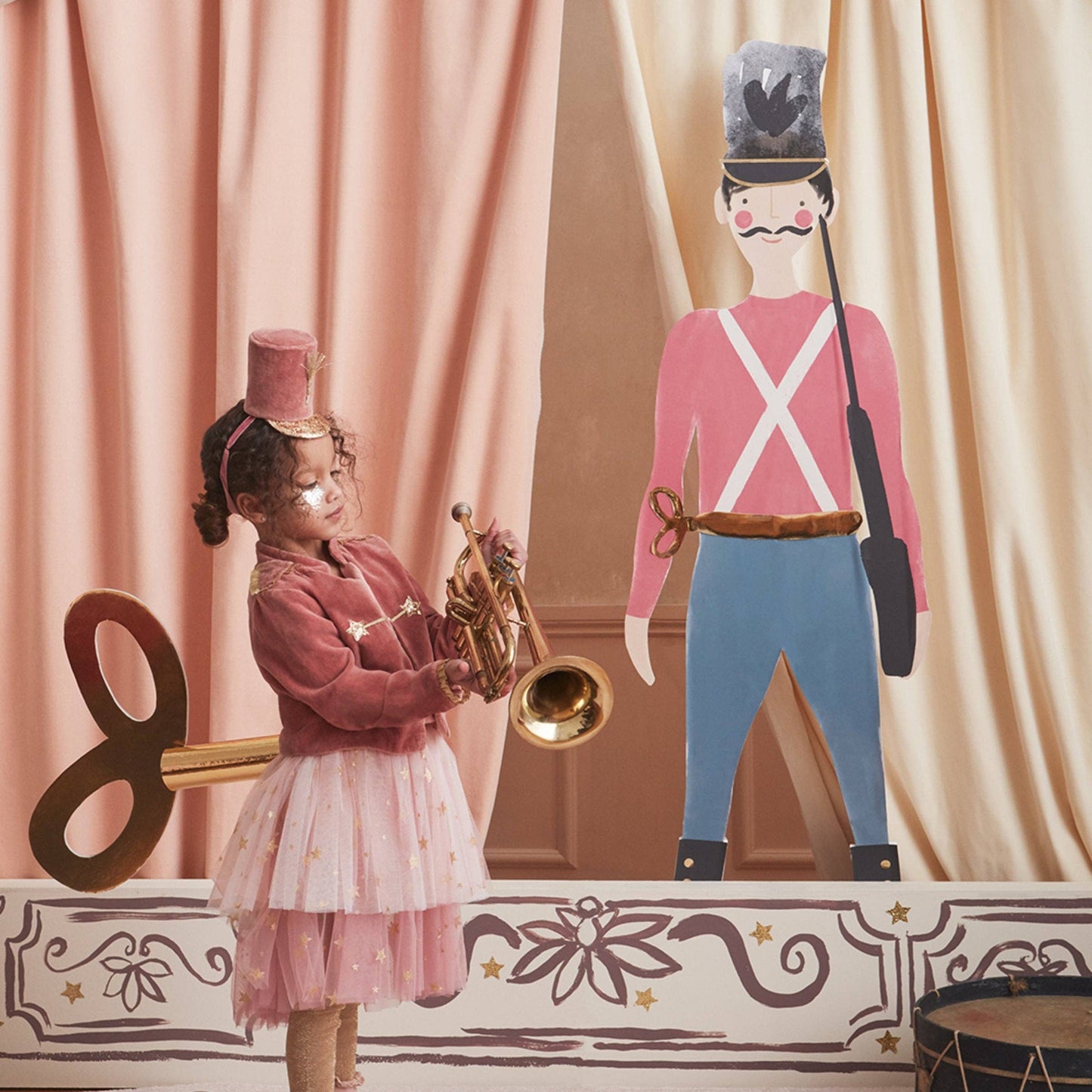 A young girl in a pink outfit stands holding a brass trumpet in front of peach curtains, with a large cardboard toy soldier and decorative props on a stage-like set.