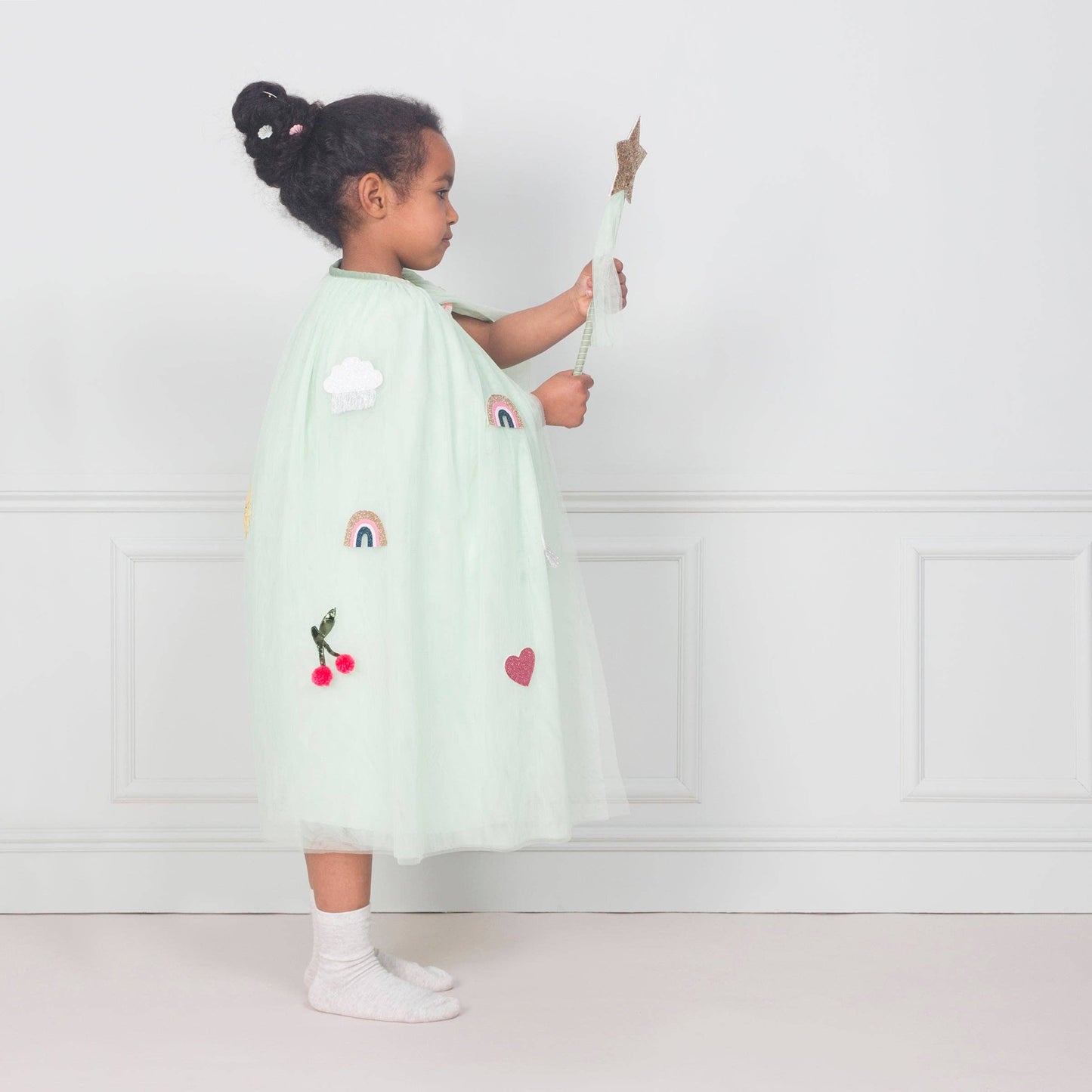 Child wearing a light green tulle cape with rainbow and heart designs against a white wall.