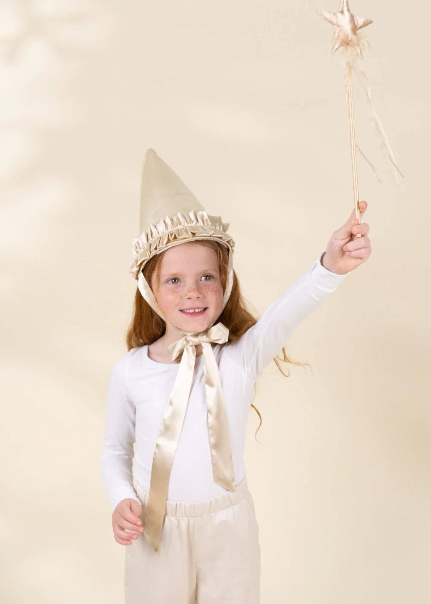 A young girl wearing a beige wizard or fairy costume with a pointed hat, white long-sleeve top, and cream pants, holding a star wand against a soft beige studio background.