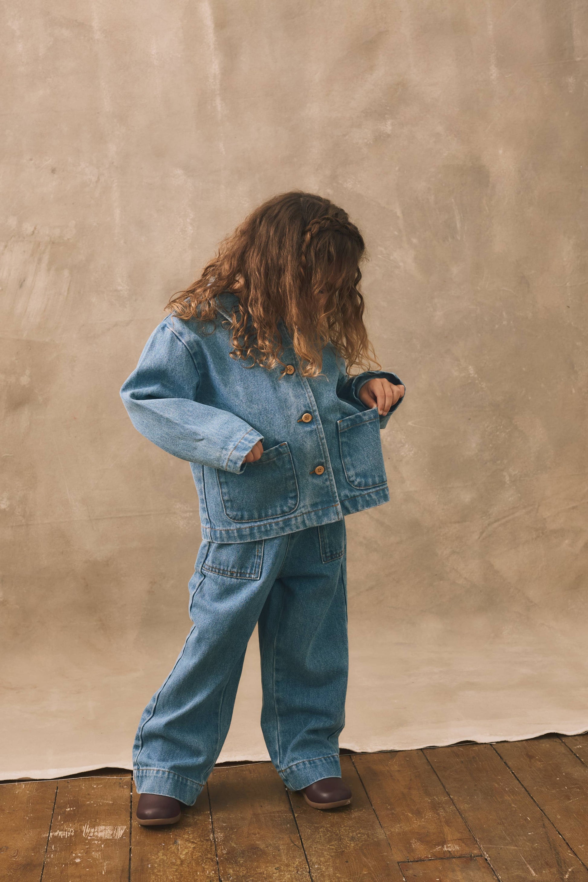 Child wearing a denim outfit standing against a beige wall.