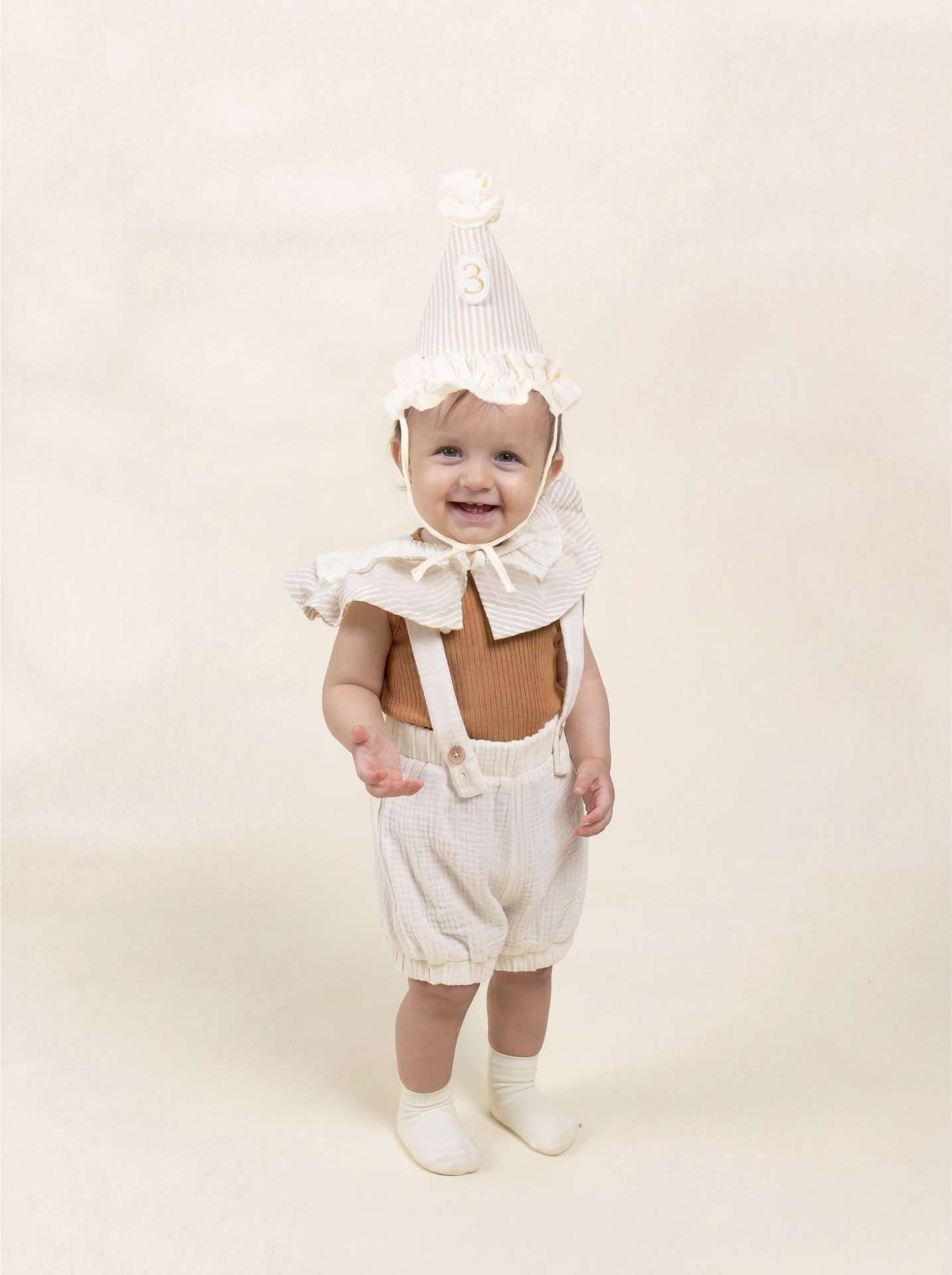 Baby wearing a white costume with a hood and suspenders on a beige background