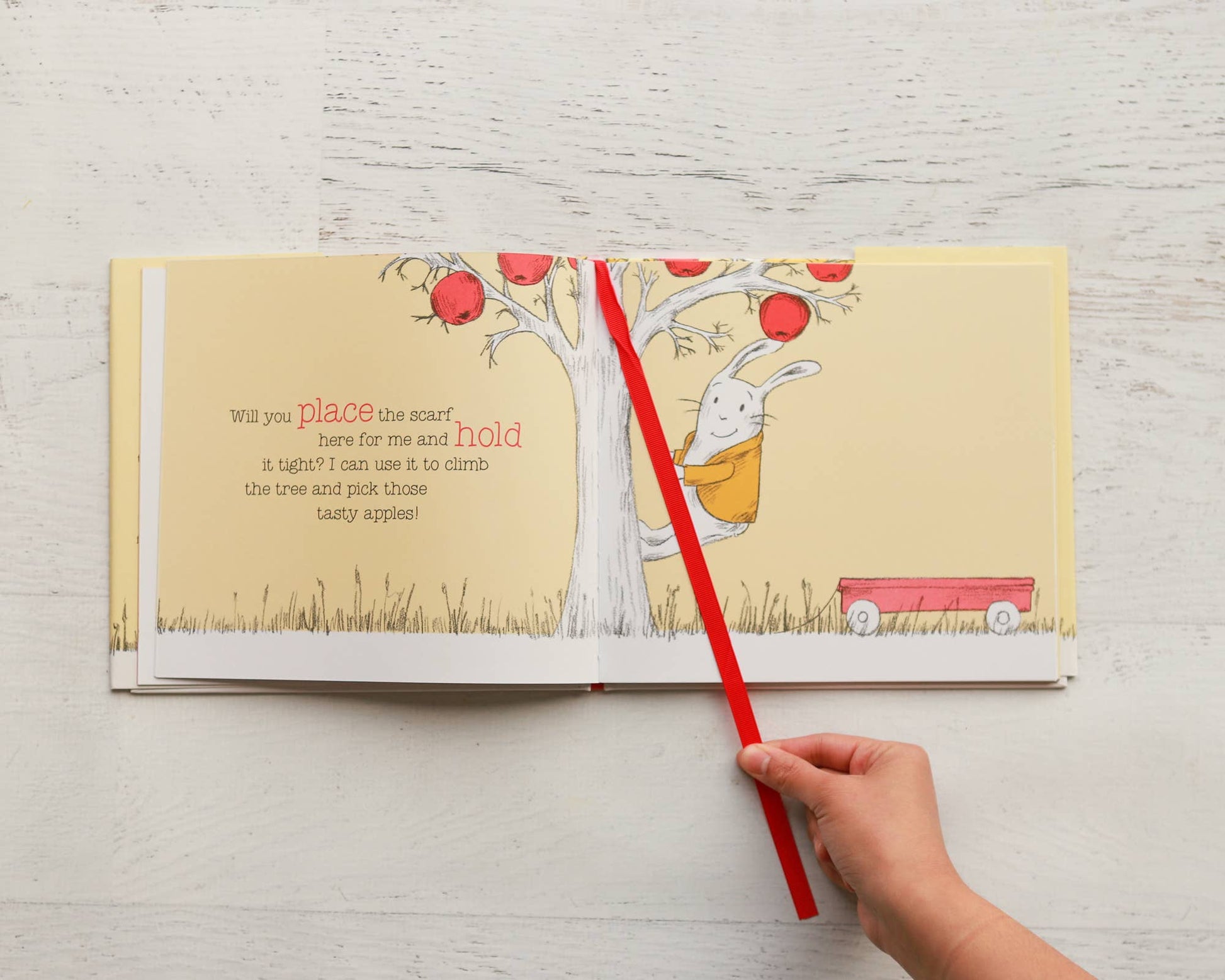 Open book with a child's hand holding a red stick, illustration of a rabbit and tree on a white wooden surface