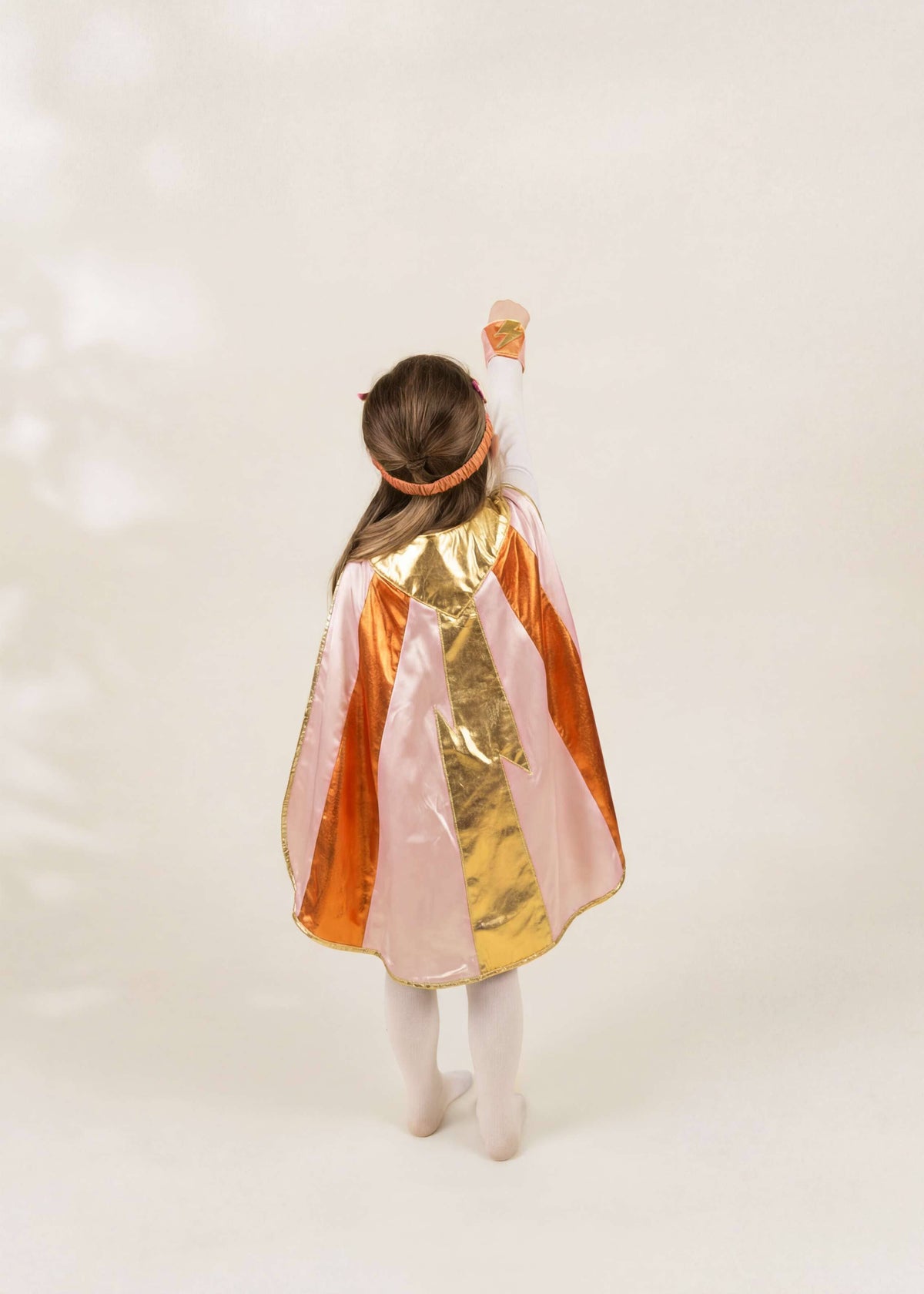 A child viewed from behind wearing a shiny costume cape with vertical panels of orange, gold, and pale pink, along with white tights and a matching headband, against a plain light background.