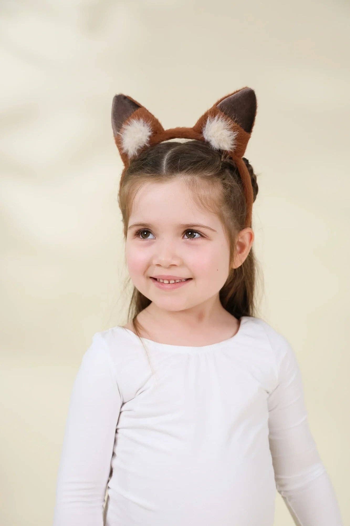 A young girl with brown hair wearing a brown and white faux fur fox ears headband, smiling against a soft beige background.