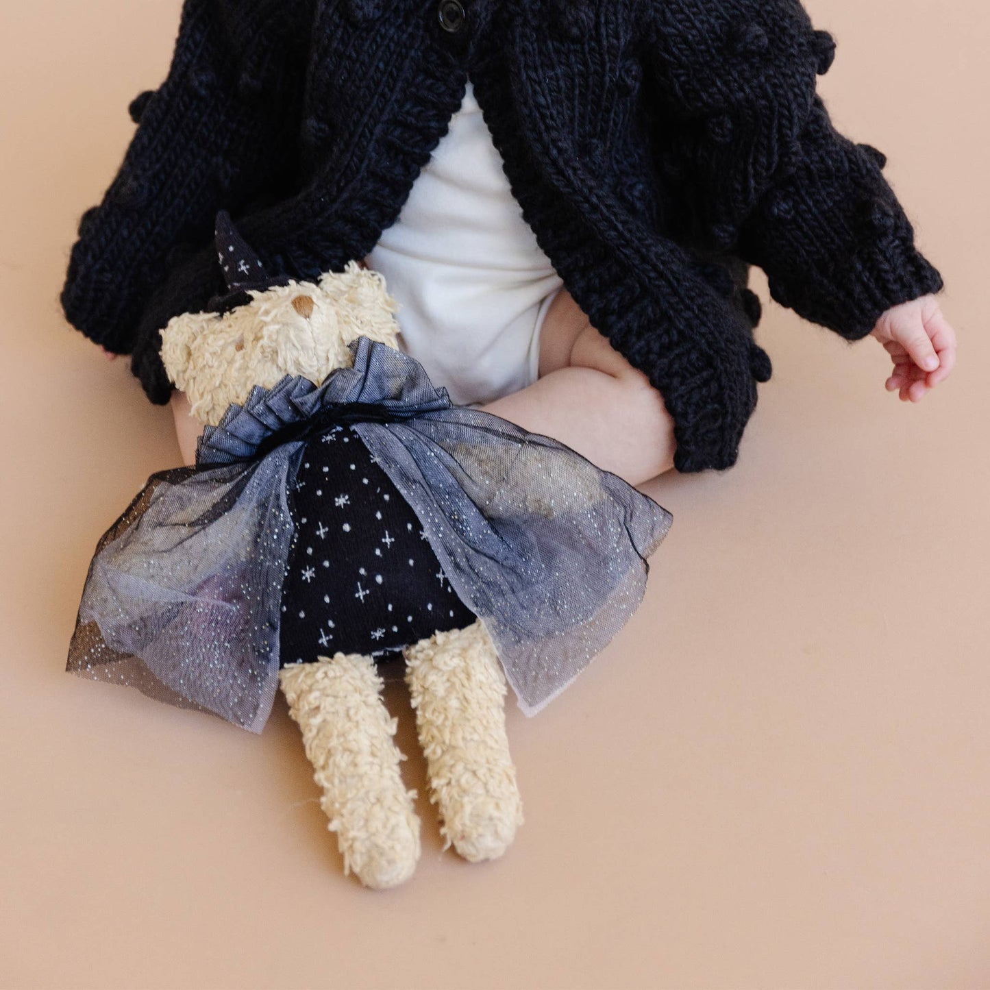 Child holding a teddy bear dressed in a black outfit with a blue bow on a beige background