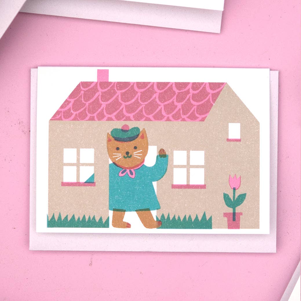Card on a pink background. The card has a lithograph print of a cat waving in front of a house.