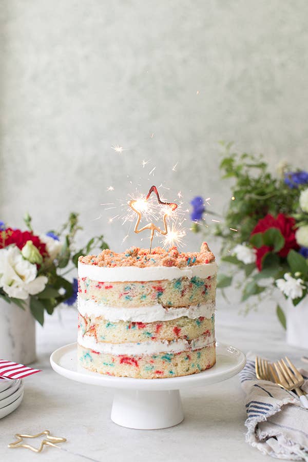 Colorful cake with star-shaped candles on a white cake stand, surrounded by flowers and a striped napkin.