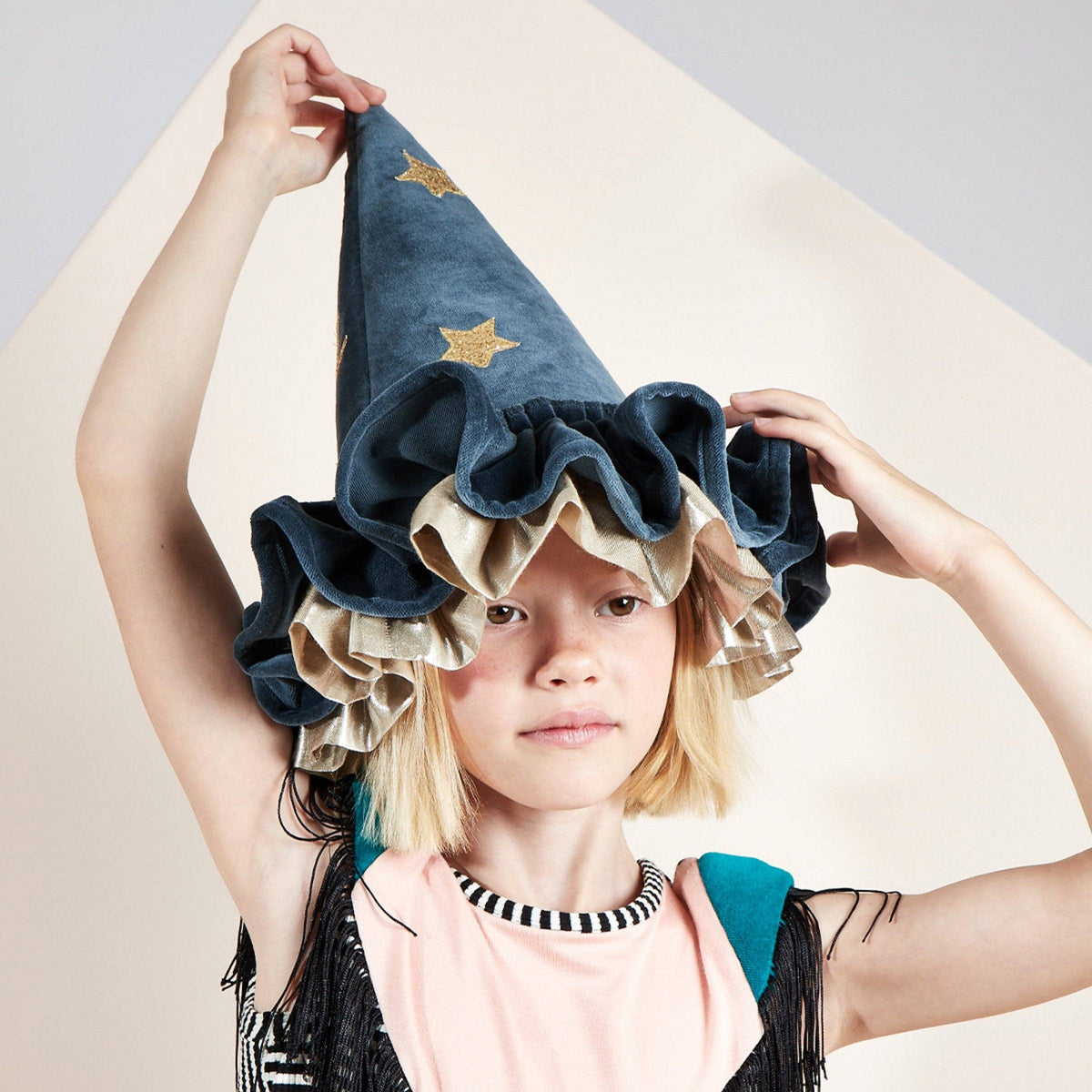 A child wearing a tall navy blue velvet wizard hat with gold star details and layered beige ruffles, posed against a minimalist beige and gray background.