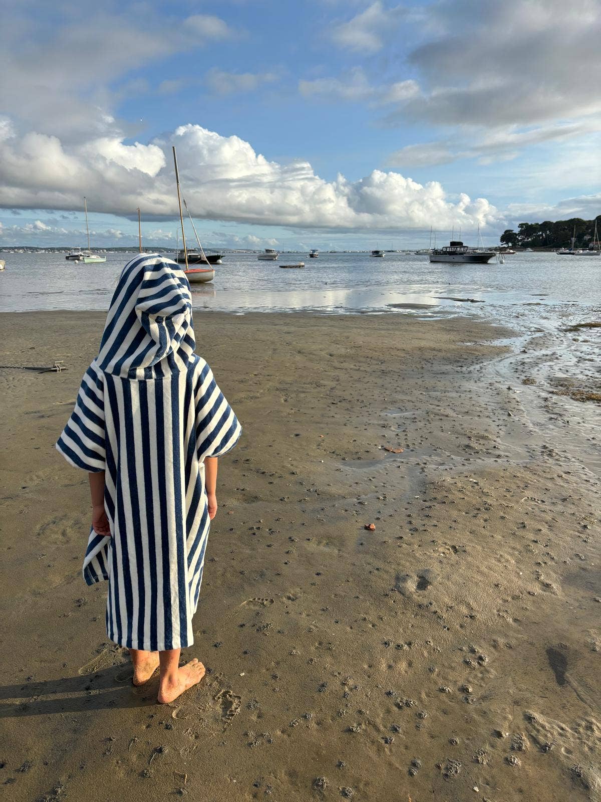 Person wearing a striped hooded towel on a beach with boats in the background