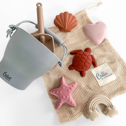 Children's sand playset with a blue pail, brown shovel, red turtle, pink starfish, and heart-, shell- and rainbow- shaped molds on a cotton mesh bag.