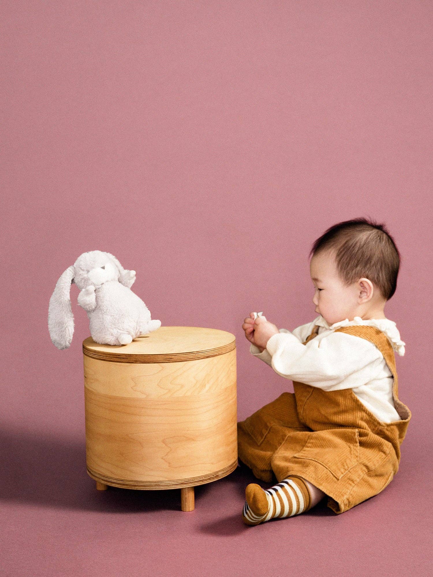 Child playing with a plush toy on a wooden stool against a pink background