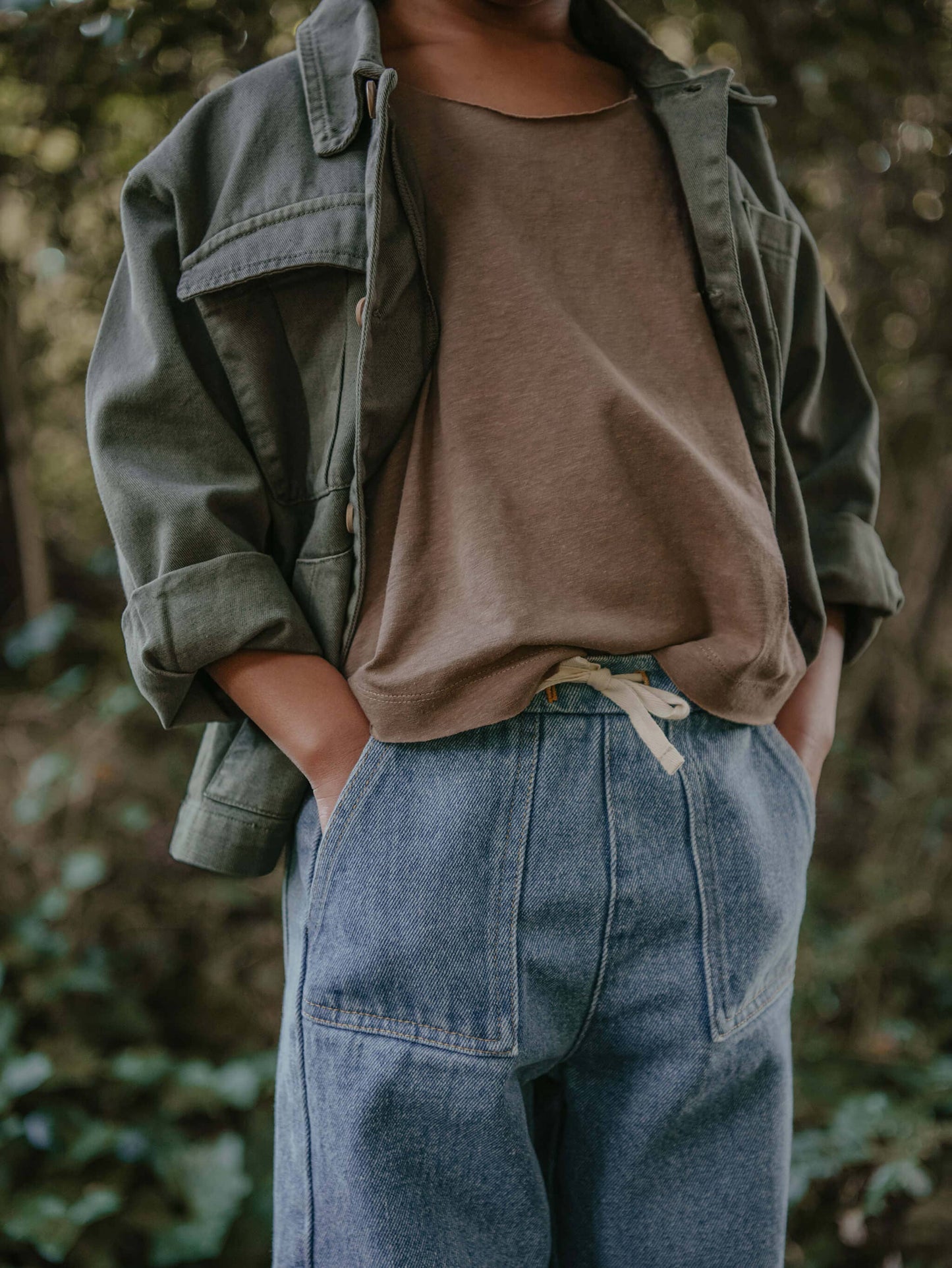 Child wearing a green jacket, brown shirt, and blue jeans standing in a forest.