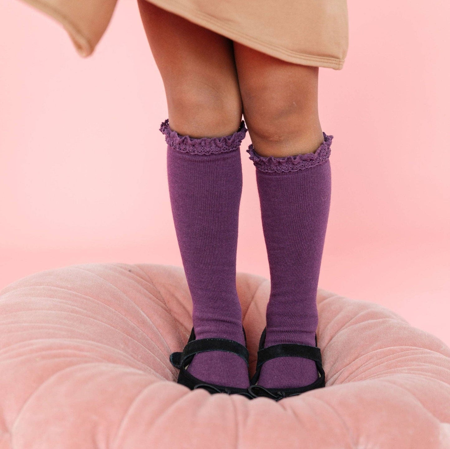 Purple knee-high socks with ruffles worn by a person on a pink cushion against a pink background