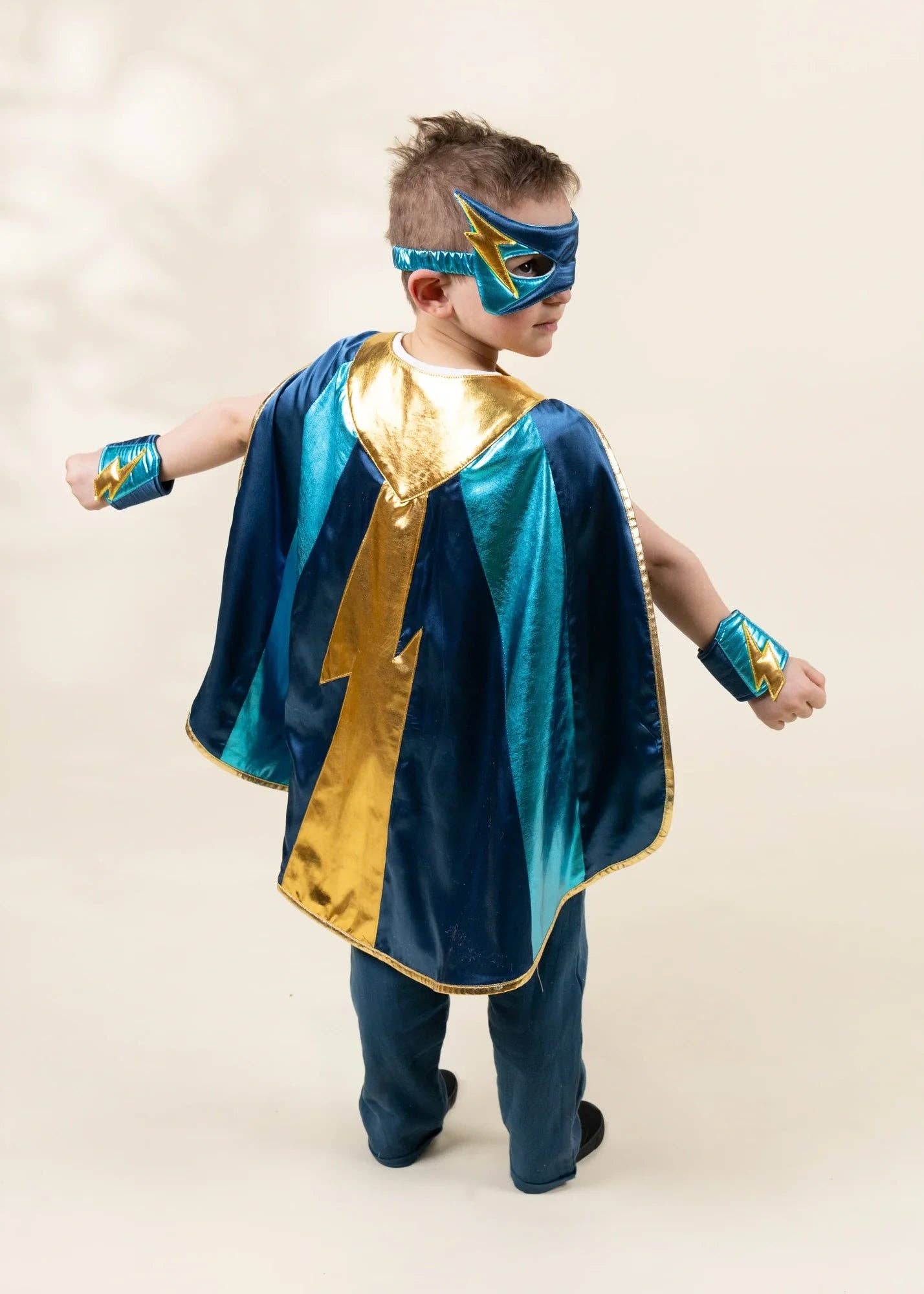 A young boy wearing a superhero costume with a blue and gold cape, matching mask, and wristbands, standing against a plain beige background.