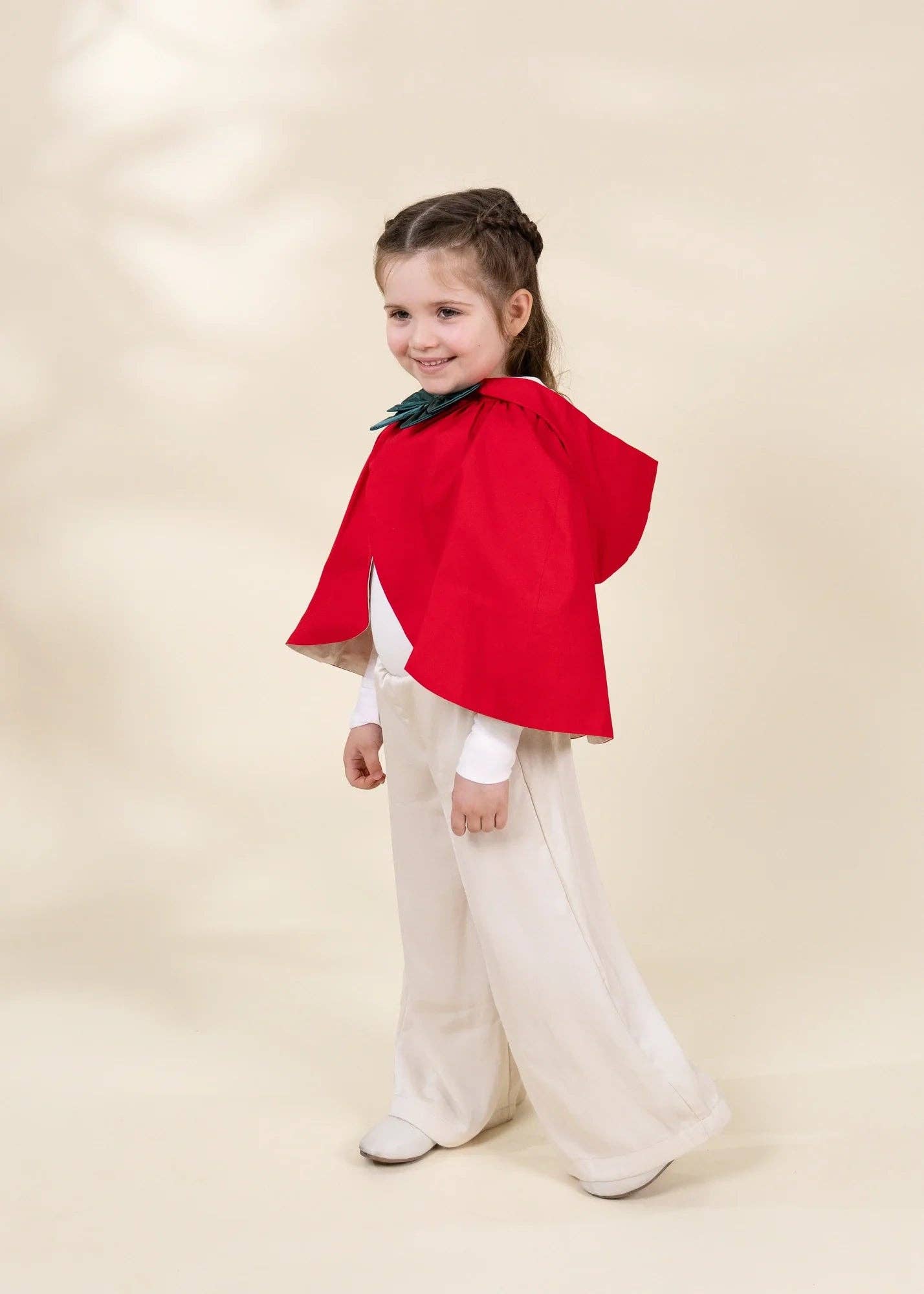 A young girl modeling a festive costume consisting of a bright red cape with a green collar, a white long-sleeve shirt, and wide-leg cream pants, set against a warm beige studio backdrop.