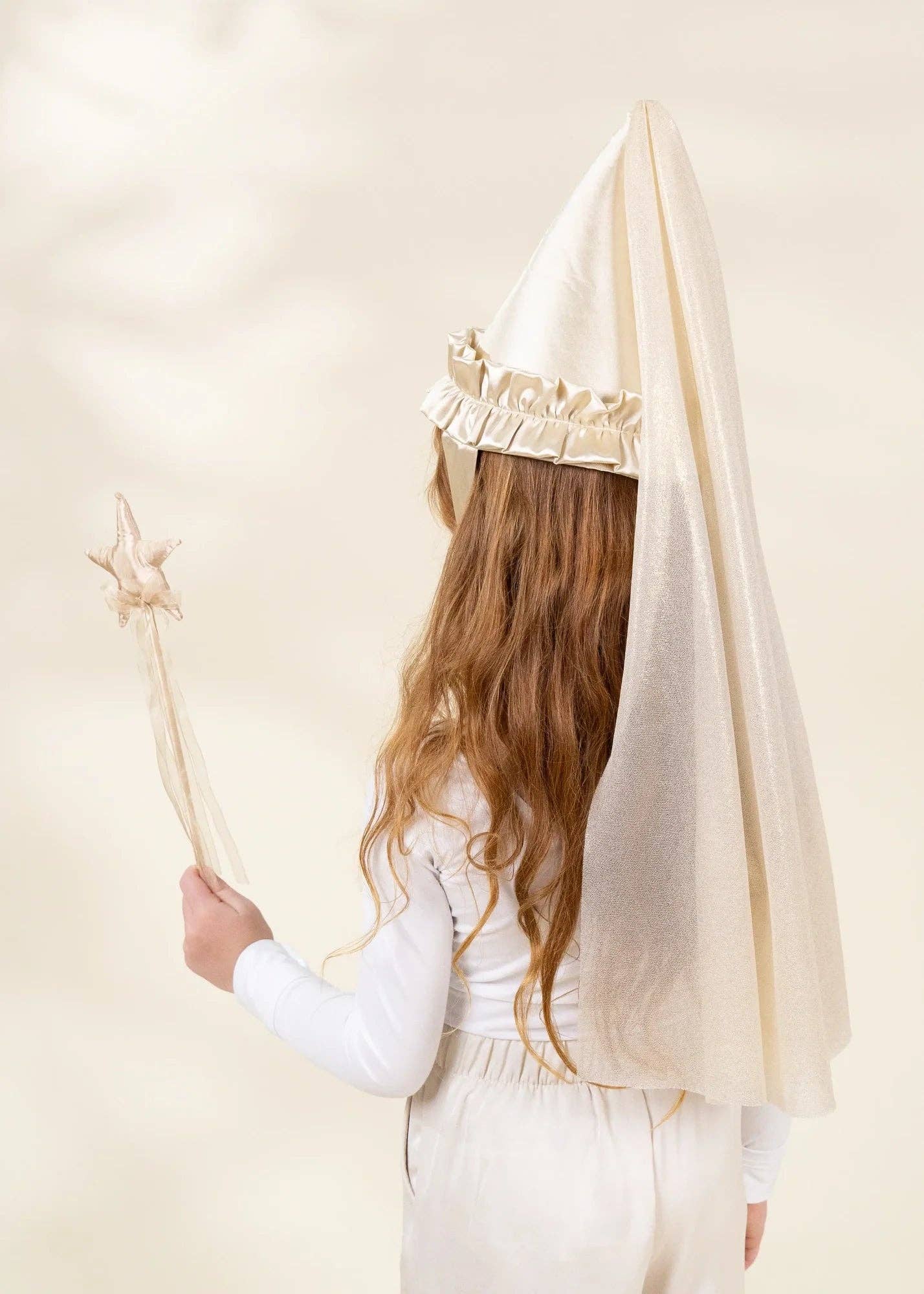 A child seen from behind wearing a cream-colored pointed hat with a ruffled trim and a long wavy light brown hair, holding a matching wand against a soft beige background.