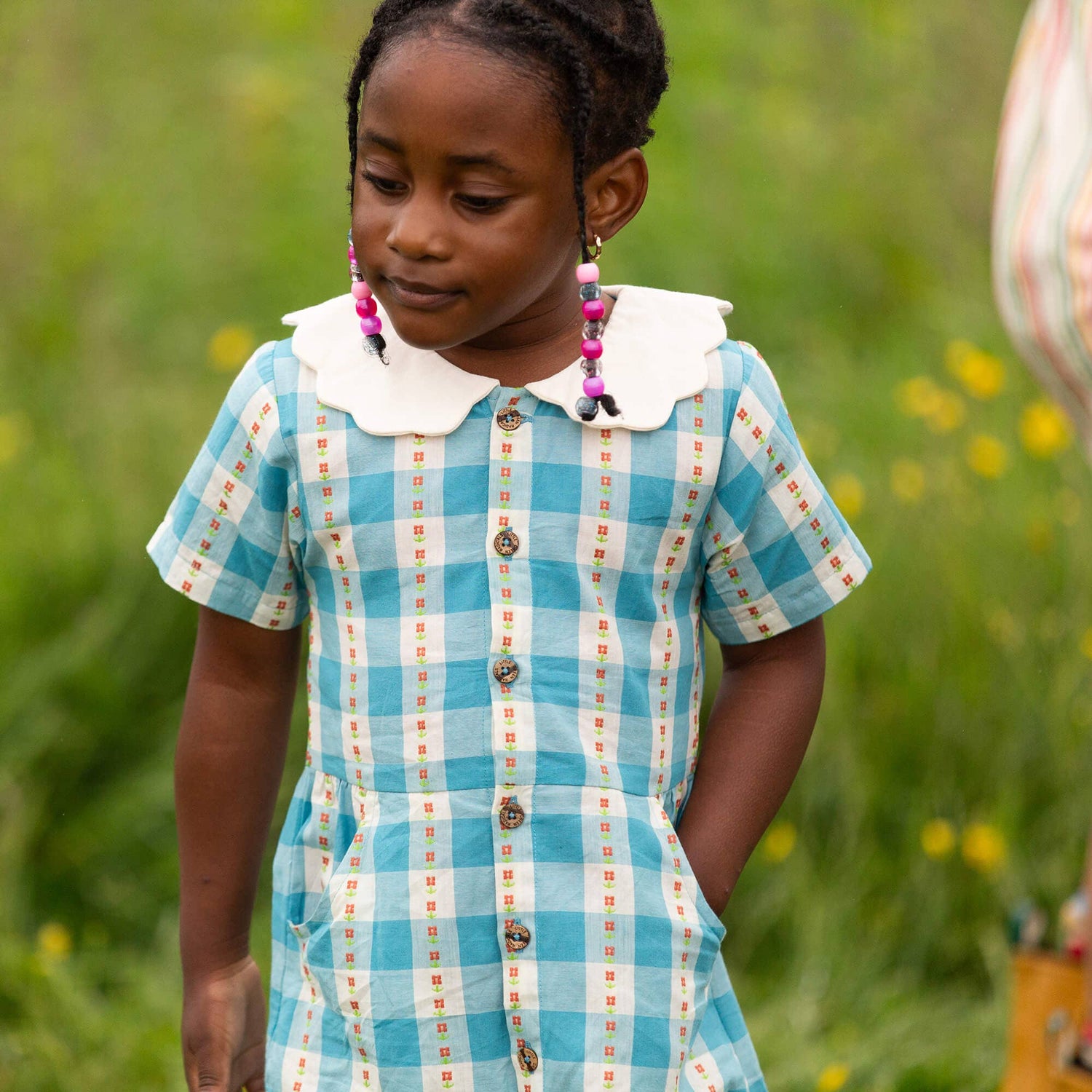 Young girl in a blue checkered dress standing in a grassy field with flowers.