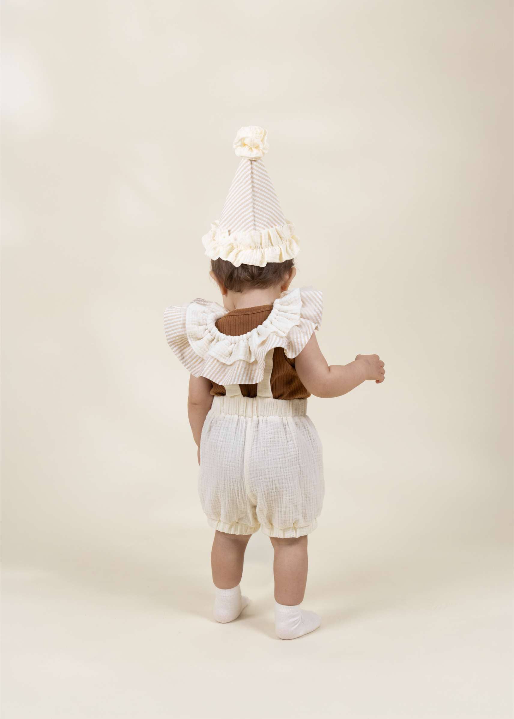 A baby viewed from behind wearing a cream and white striped party hat with a ruffled trim, a brown sleeveless top, and cream checkered bubble shorts, standing against a solid cream background.