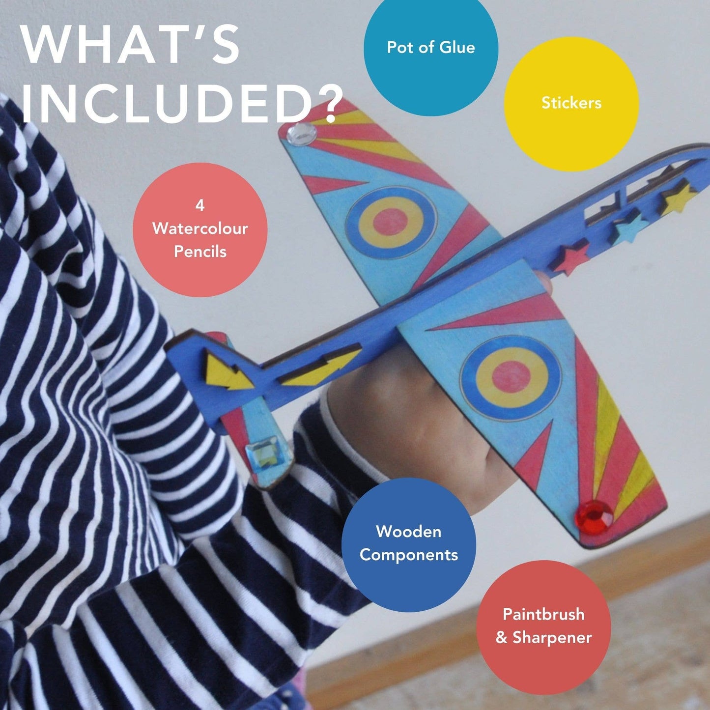 Colorful toy airplane with labeled components, held by child with striped shirt. "What's Included? 4 watercolor pencils, pot of glue, stickers, wooden components, paintbrush and sharpener"