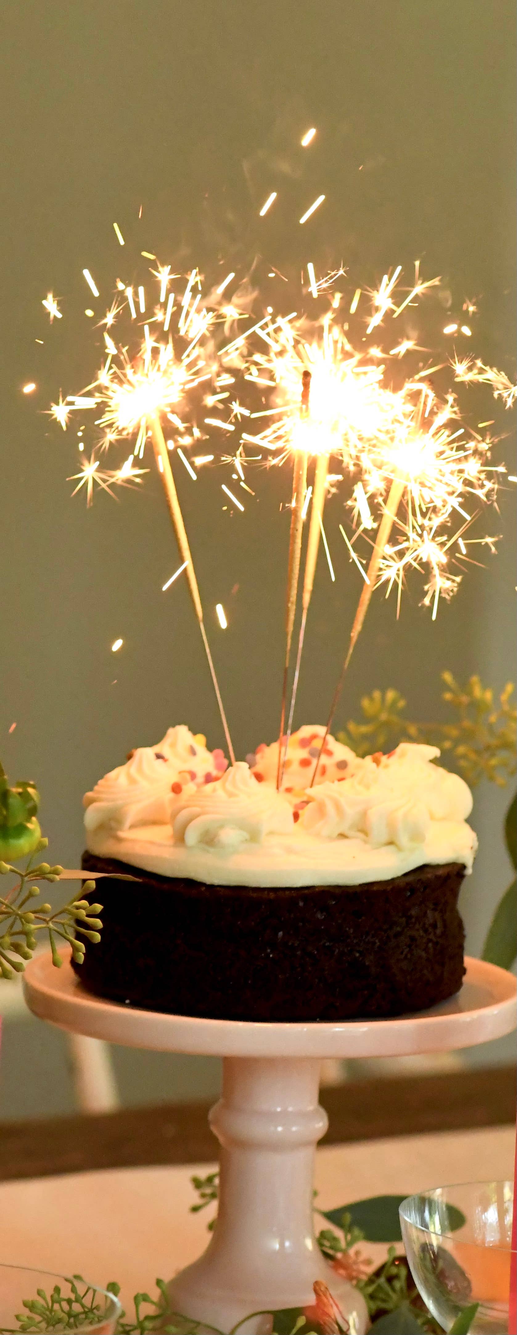 Chocolate cake with white frosting and lit sparklers on a white stand against a green background