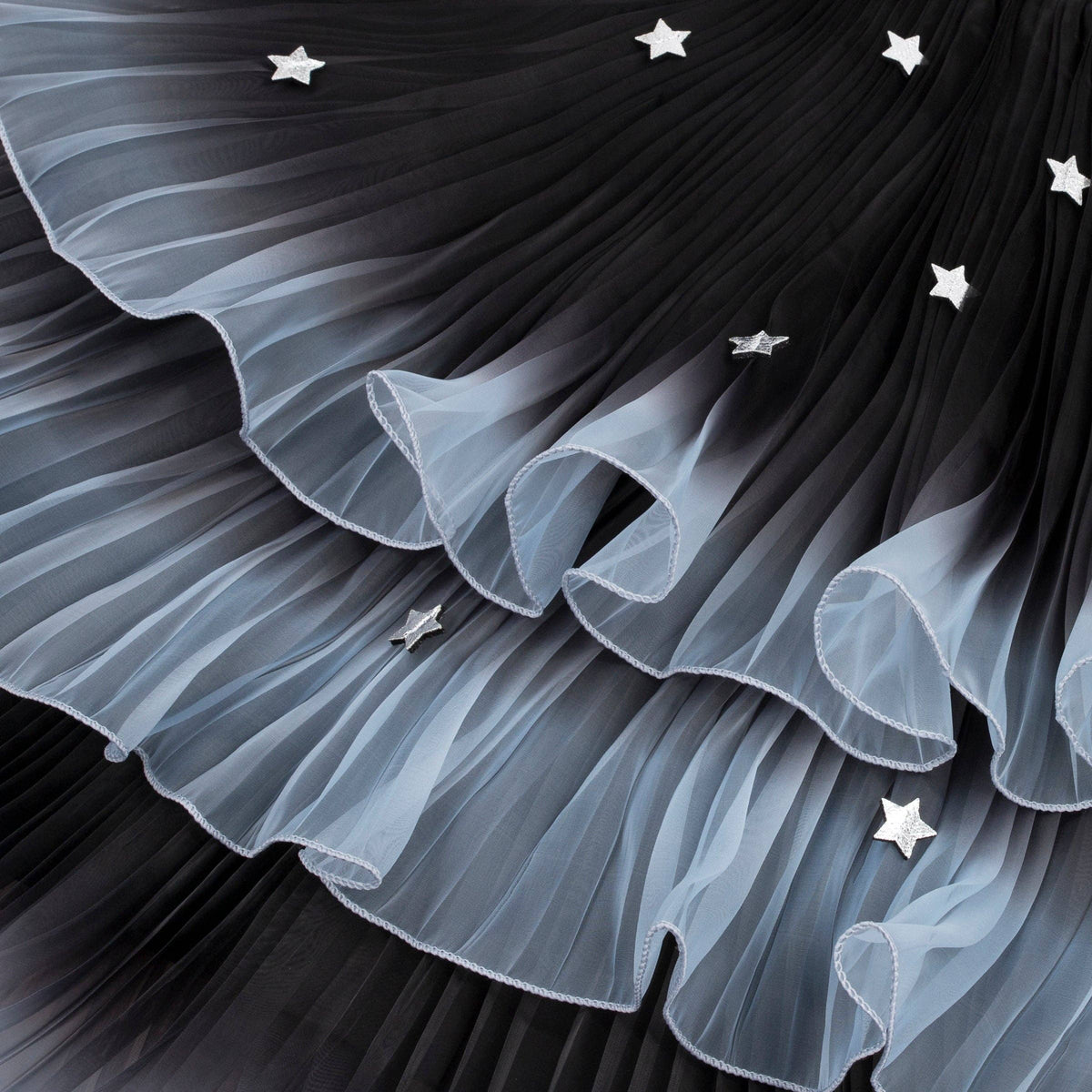 A close-up of a pleated tulle fabric, likely a skirt or dress, featuring a black-to-light-gray ombre gradient with small silver star embellishments.
