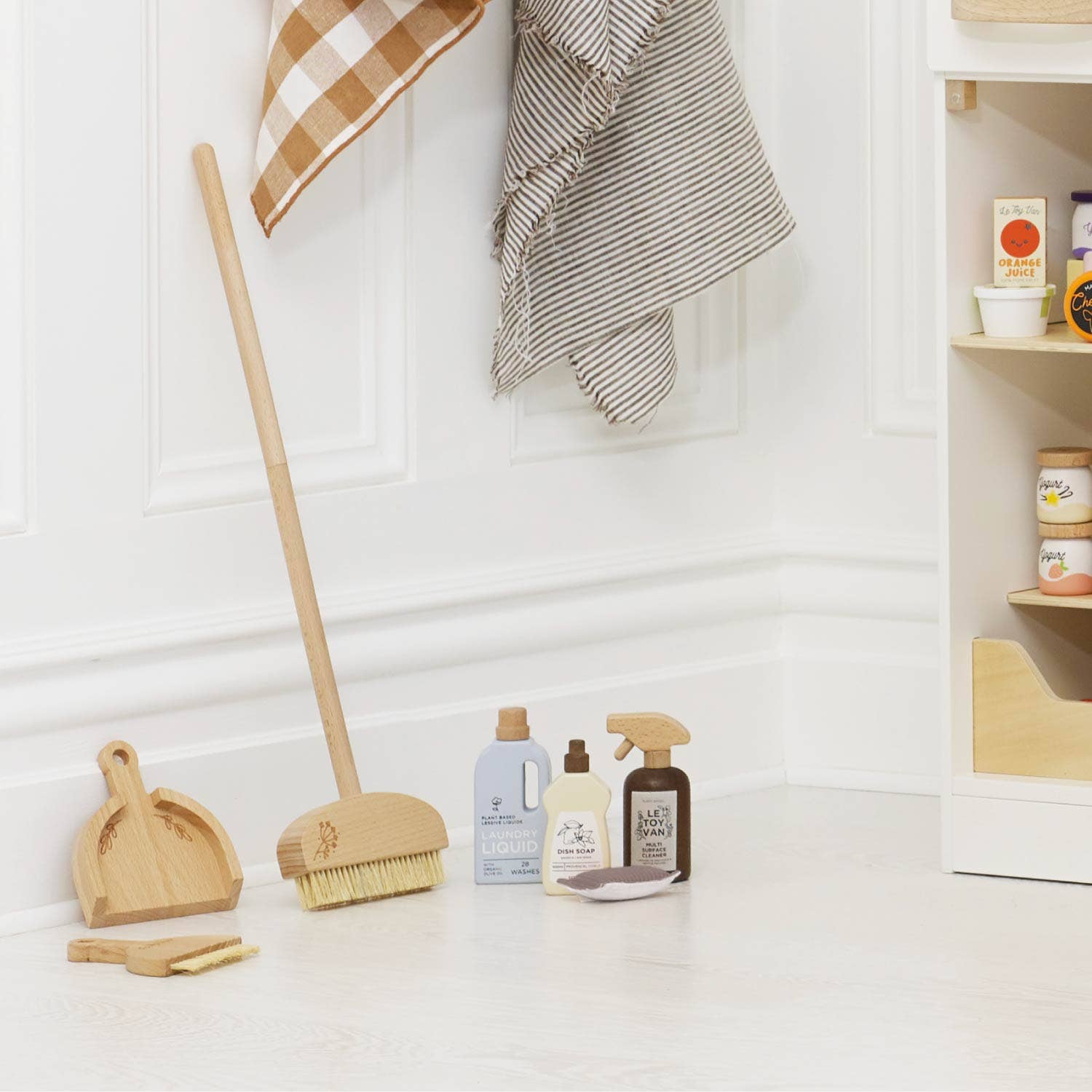 Pretend play cleaning supplies including a broom, dustpan, and cleaning bottles on a white surface with a checkered towel in the background.