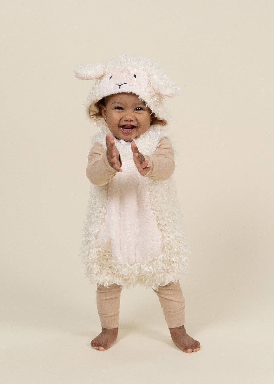 A baby wearing a fluffy white sheep costume with a hood featuring a sheep face, paired with beige leggings, standing against a solid cream background.