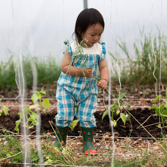 Child in a garden wearing checkered overalls and rain boots.